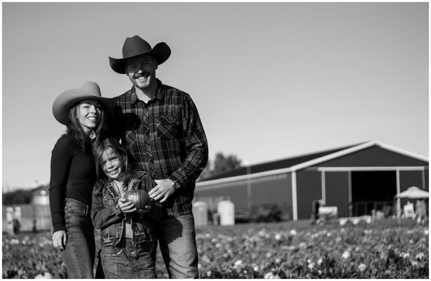 Black and white photo of a family of three in cowboy hats smiling on a farm with a red barn.
