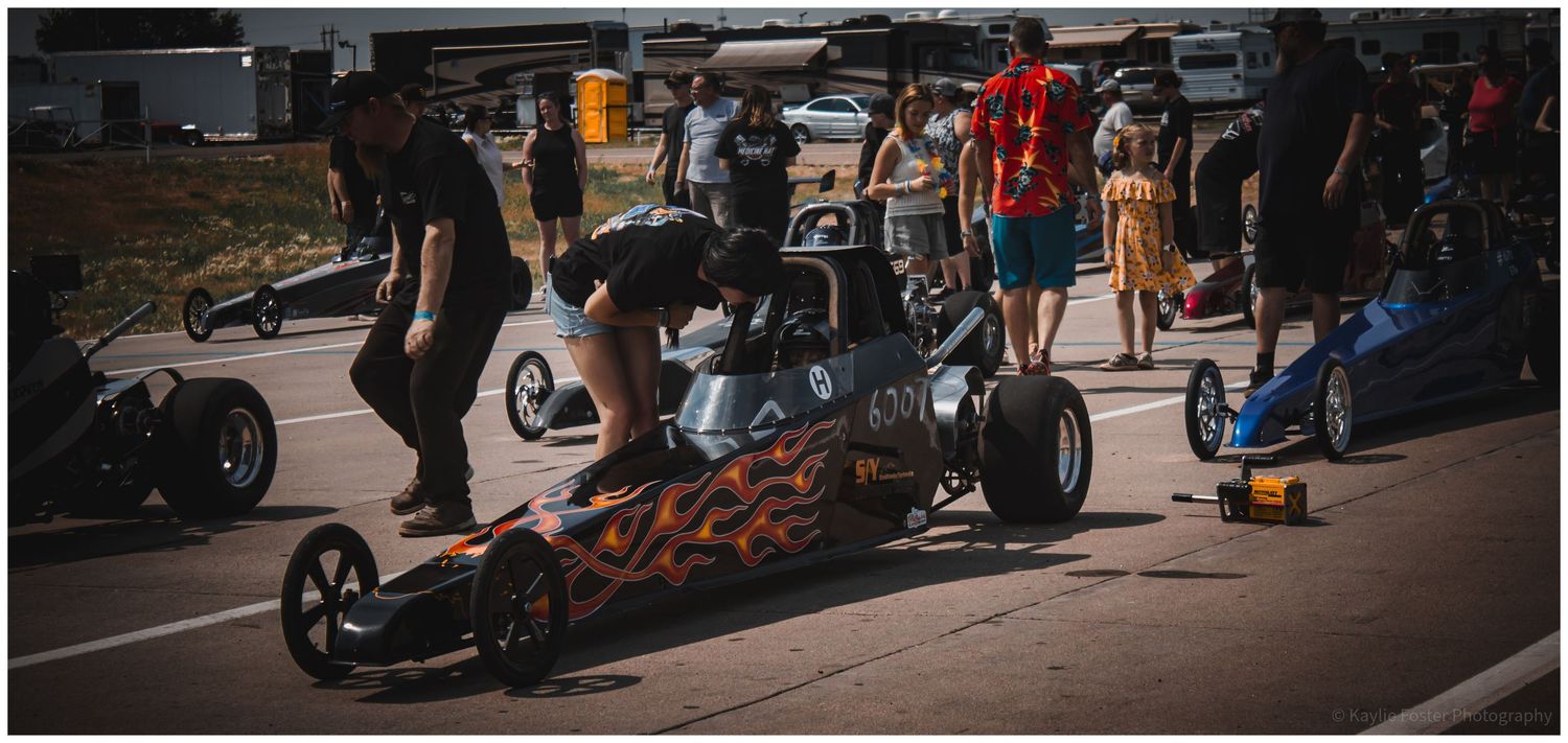 Flame-painted dragster race car being prepared by crew members at an outdoor drag racing event.