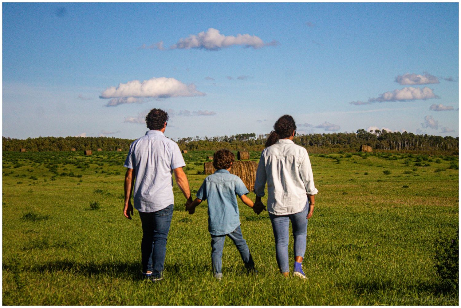 Family of three holding hands walking through a lush green meadow under a blue sky.