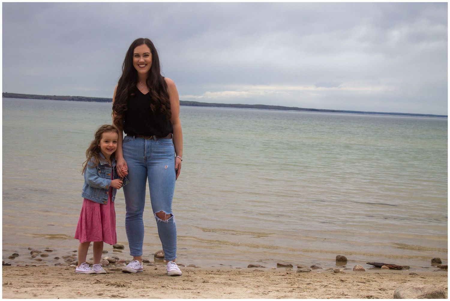 Young woman and little girl in pink dress standing on sandy beach.