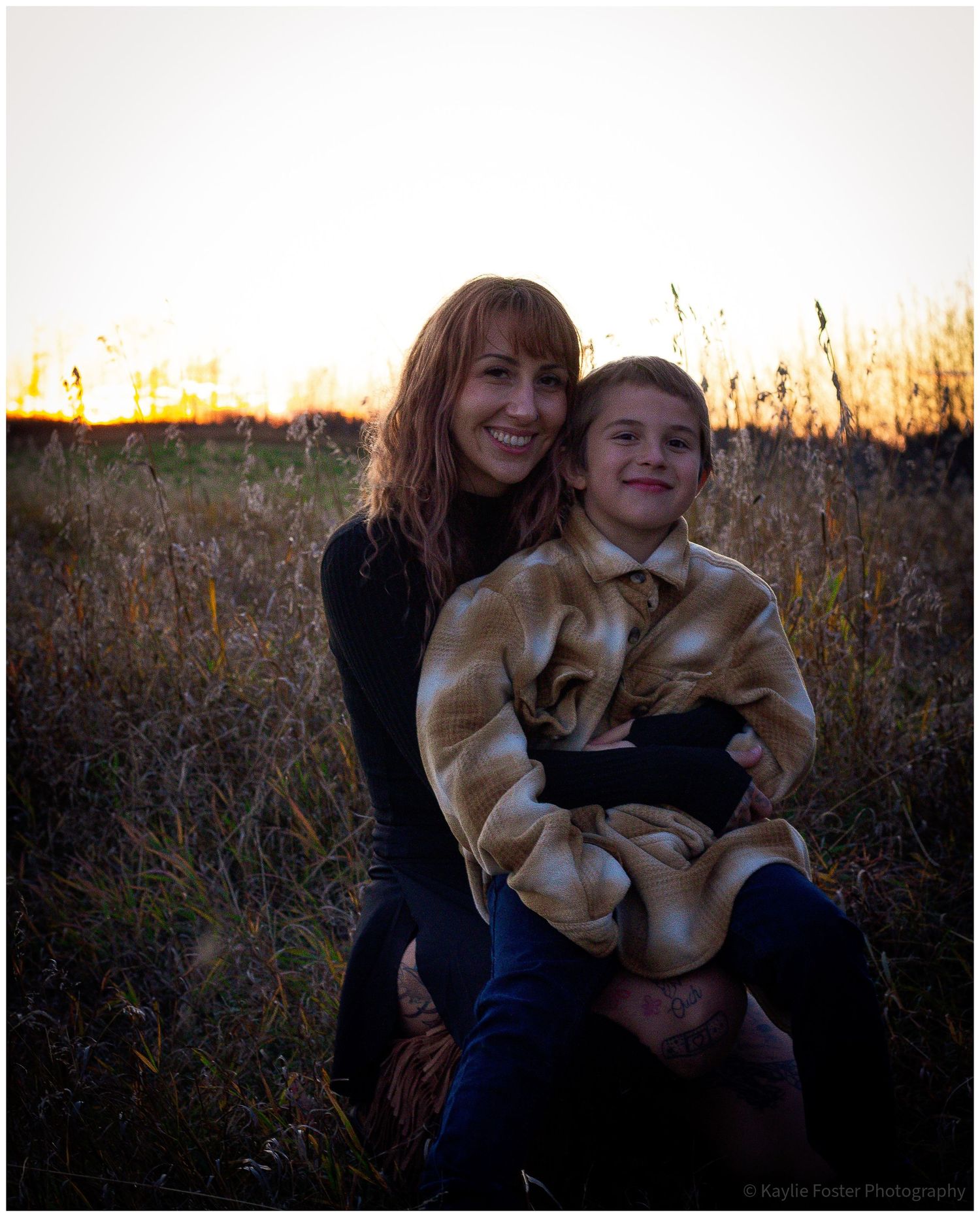 A smiling woman with red hair holds a child in a golden field at sunset during autumn.