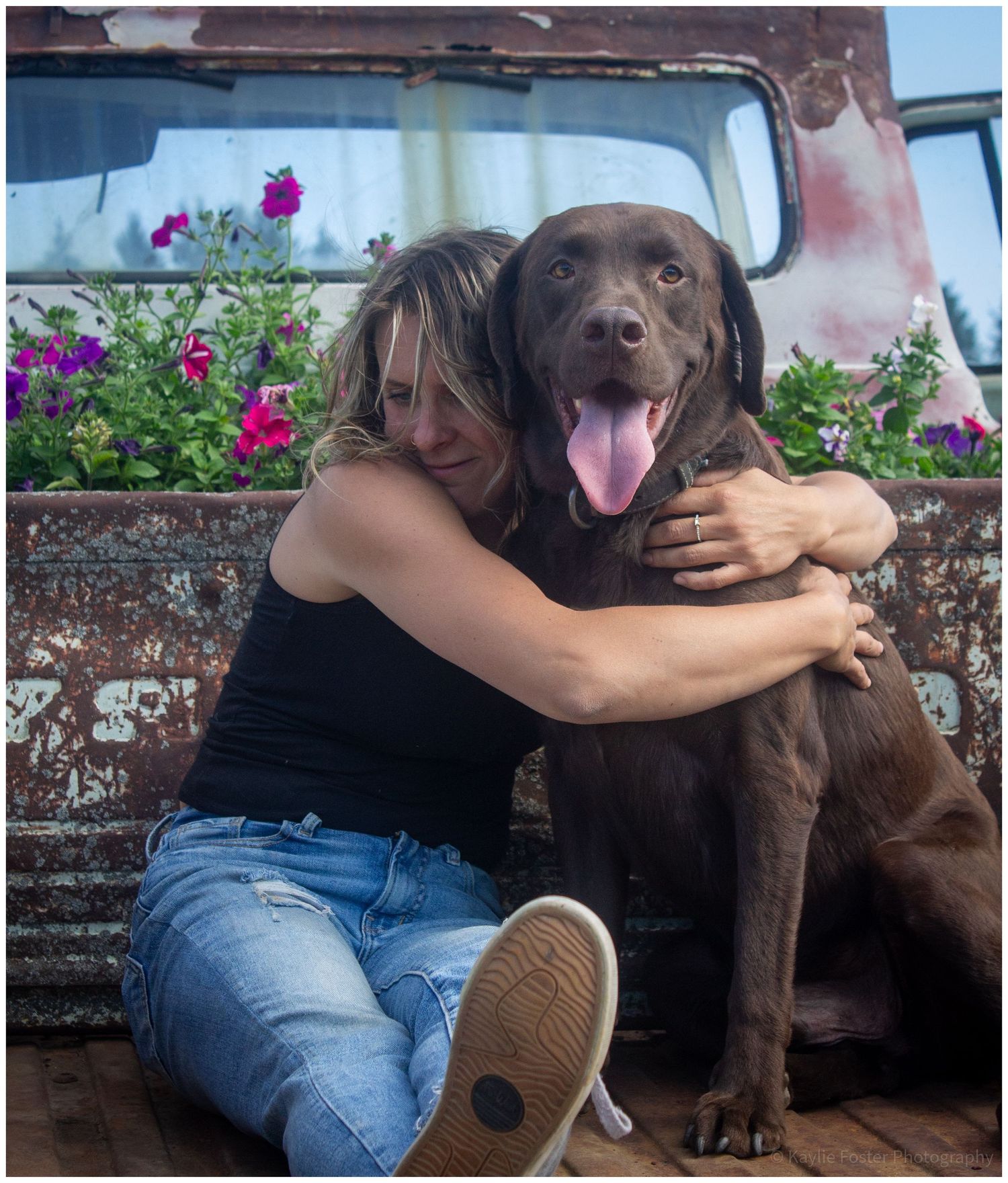 A woman joyfully embraces a chocolate Labrador beside a rusty vintage truck with purple flowers.