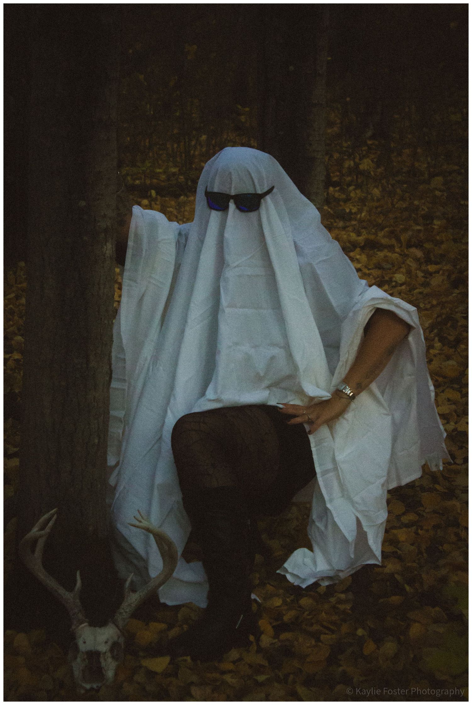 A person dressed as a ghost in a white sheet with sunglasses poses in an autumn forest beside a deer skull.