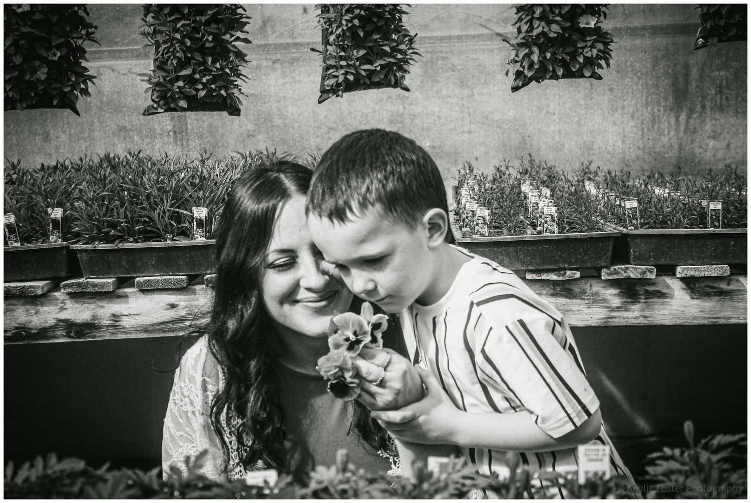 Black and white photo of a mother and young boy smelling a flower together outdoors near garden plants.