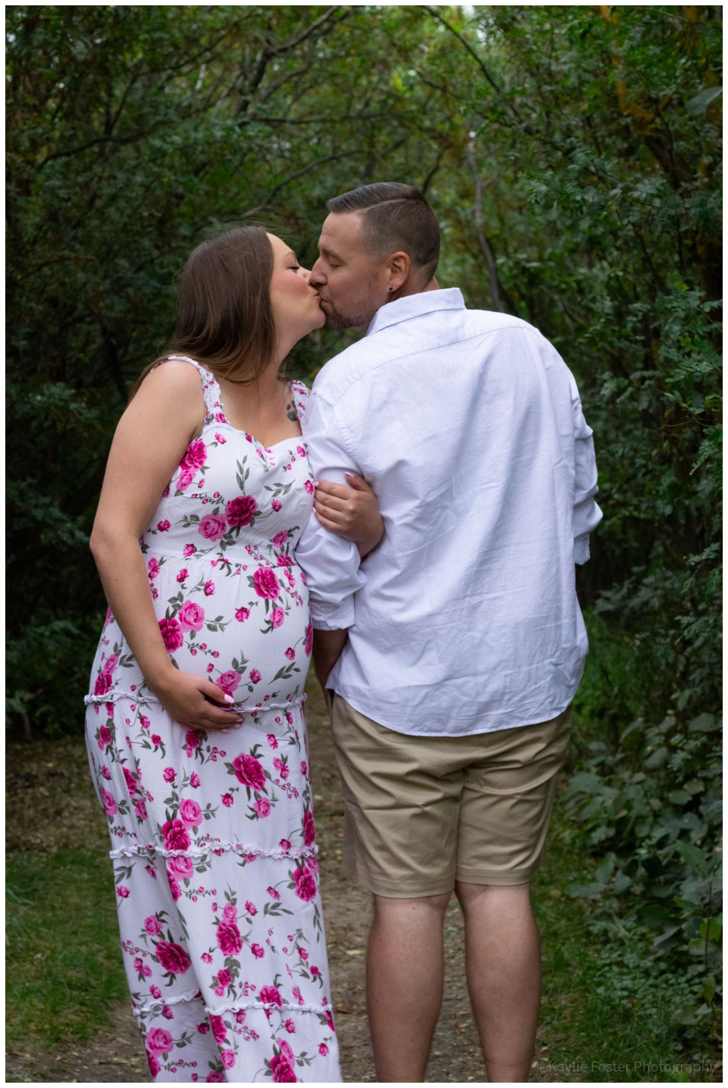 Pregnant couple sharing a kiss outdoors, woman in floral maternity dress cradling baby bump, man in white shirt and khaki shorts.