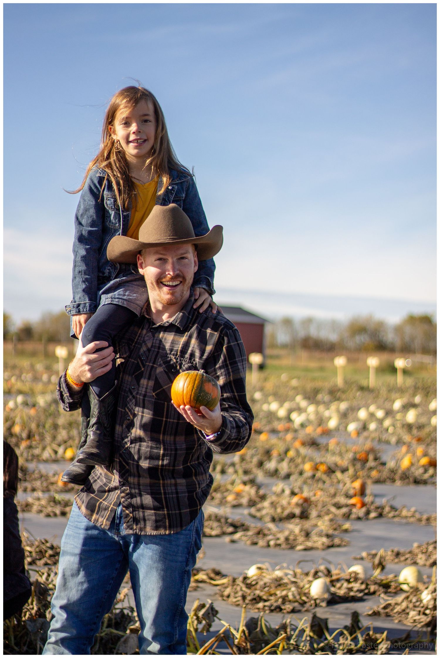 Man in cowboy hat carries girl on shoulders while holding small pumpkin in sunny pumpkin patch field.