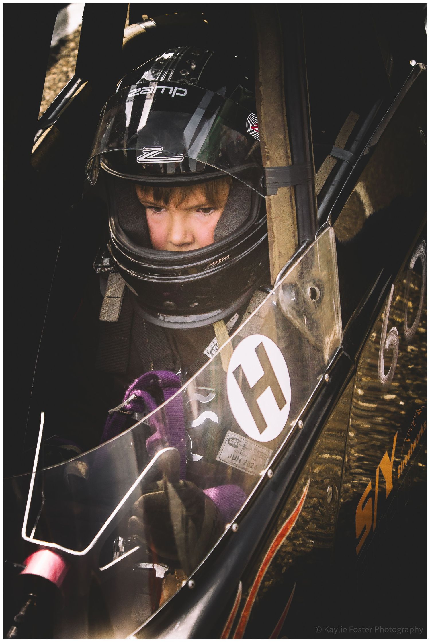 Young race car driver in a Zamp helmet seated inside a black flame-detailed dragster cockpit.