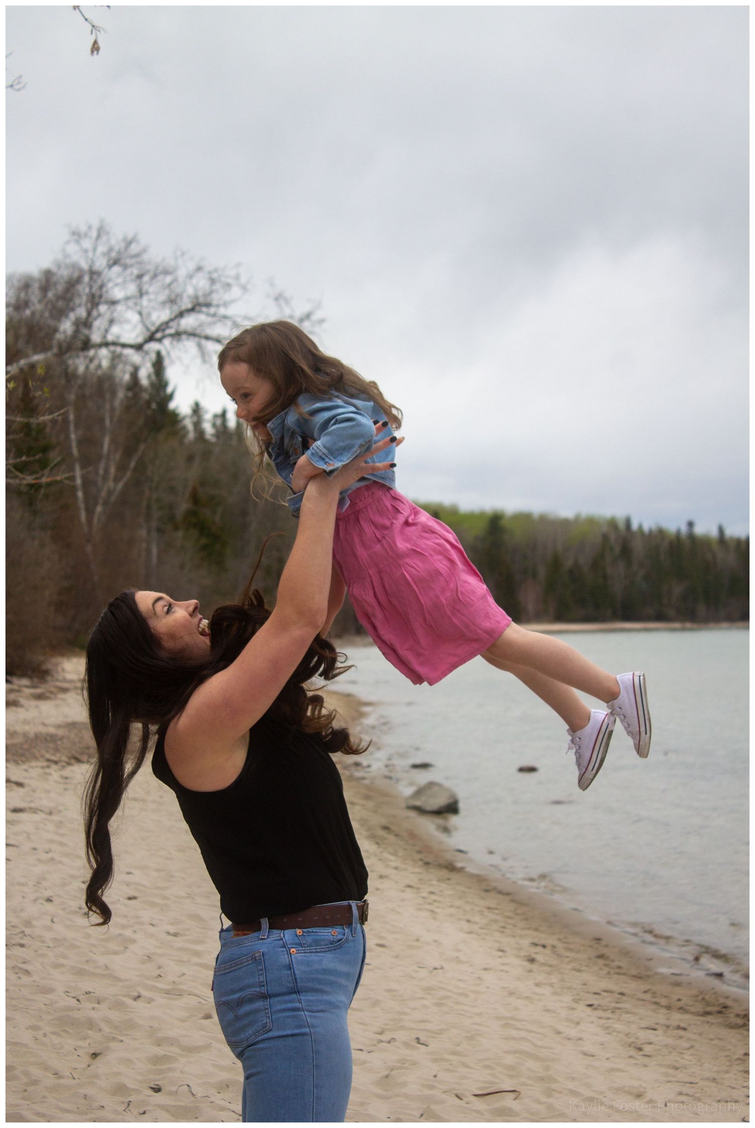 Woman lifting young girl in the air on a sandy beach by a lake.