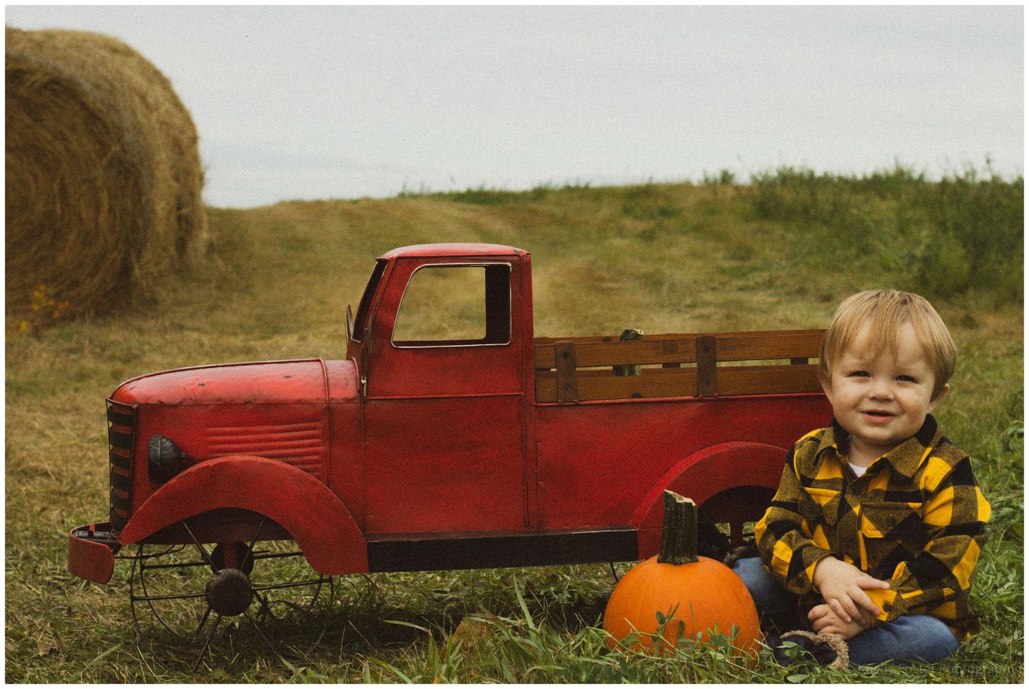 A young boy in a plaid shirt sits beside pumpkins next to a vintage red truck on a fall farm with hay bales.