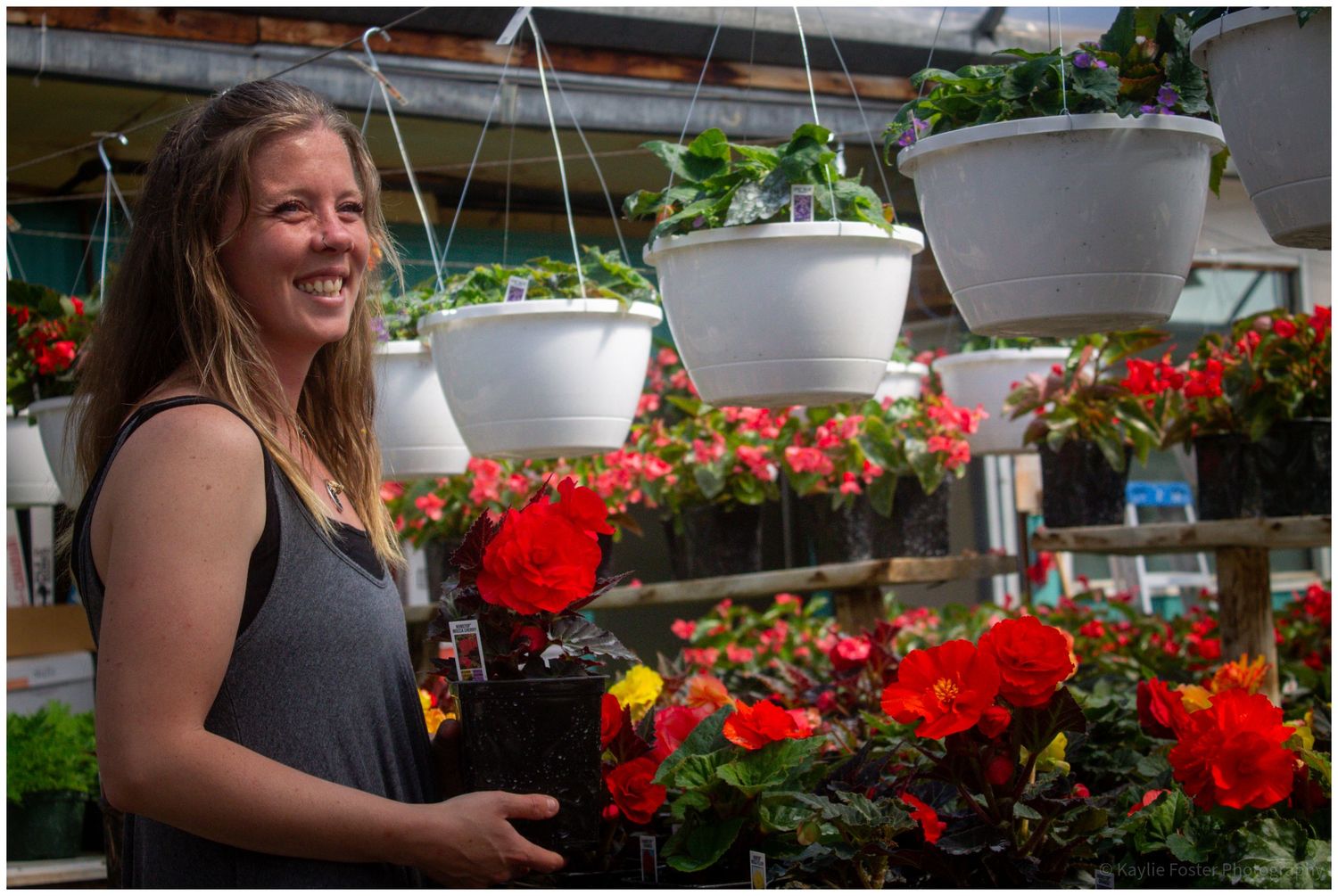 Smiling woman holding red flowers in a greenhouse with hanging white planters and colorful blooms in the background.