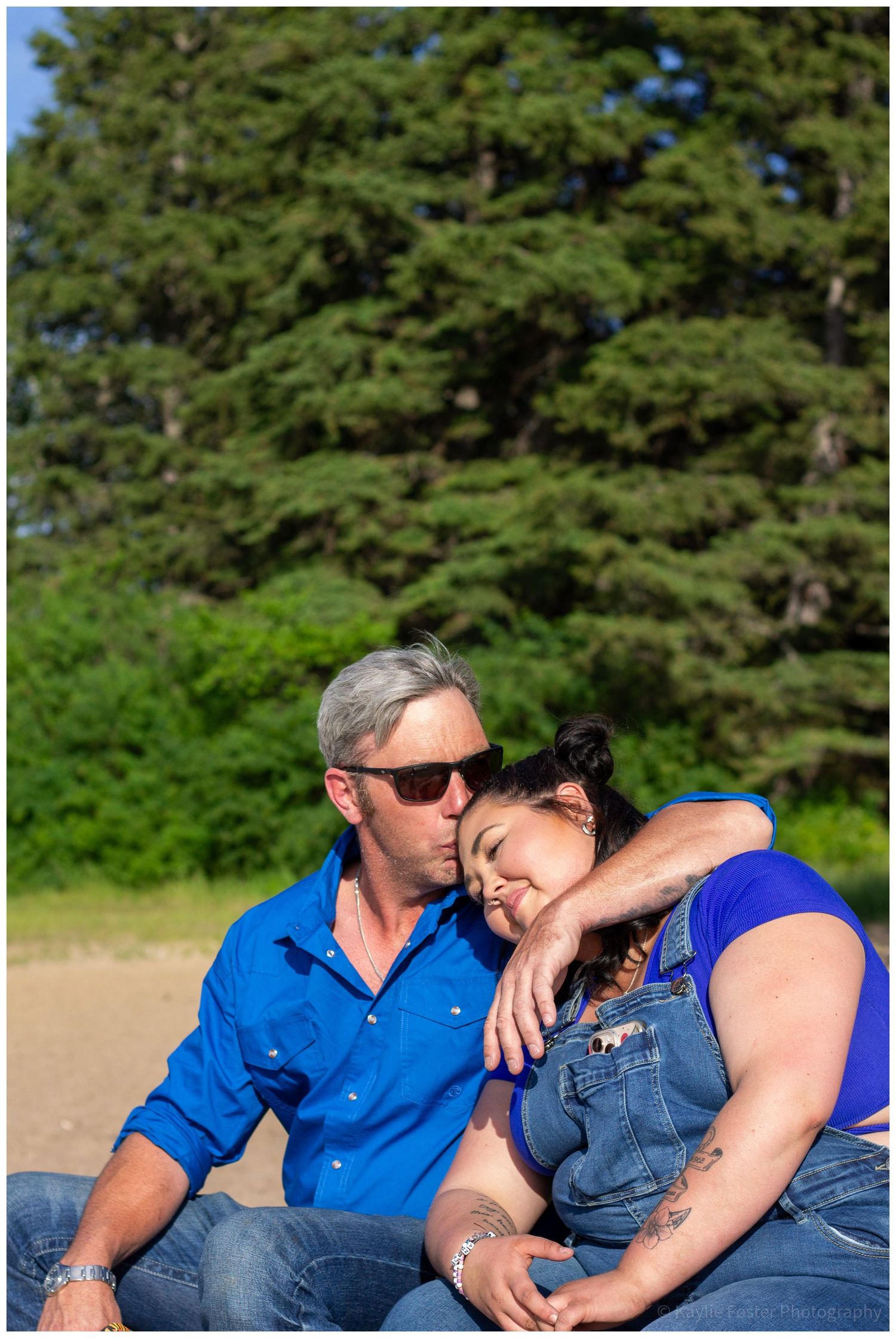 Couple in blue shirts sharing a tender moment outdoors near pine trees.