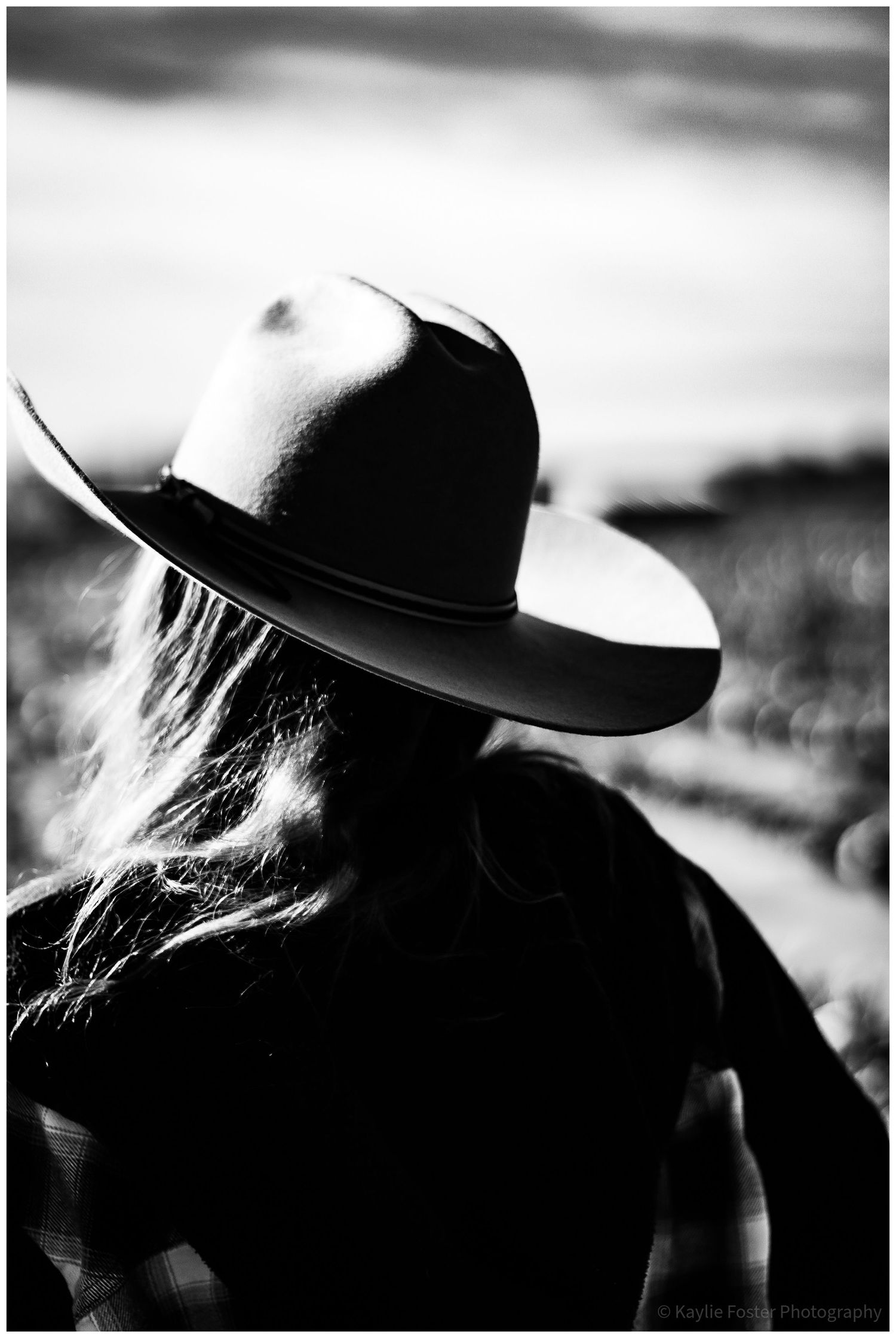 Black and white photo of a woman with long hair wearing a wide-brim cowboy hat, viewed from behind.