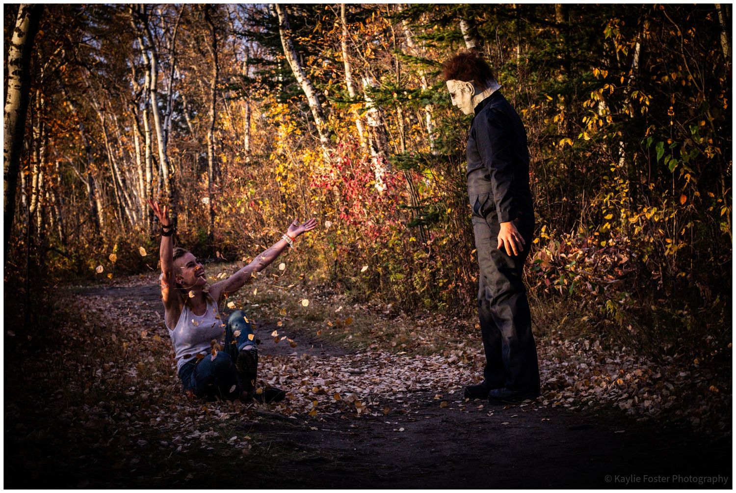 A woman reaches out toward a masked figure on a forest path surrounded by autumn foliage in a dramatic Halloween scene.
