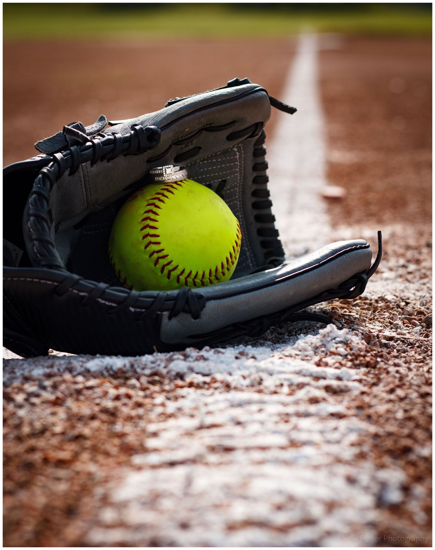 A bright yellow softball rests inside a black leather glove on a dirt baseball field near the chalk foul line.