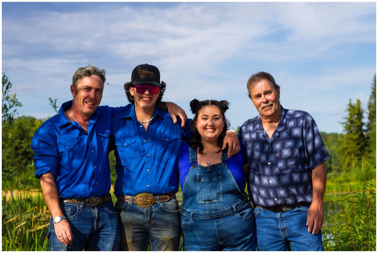 Four people in blue shirts smile together outdoors on a sunny day with green trees in the background.