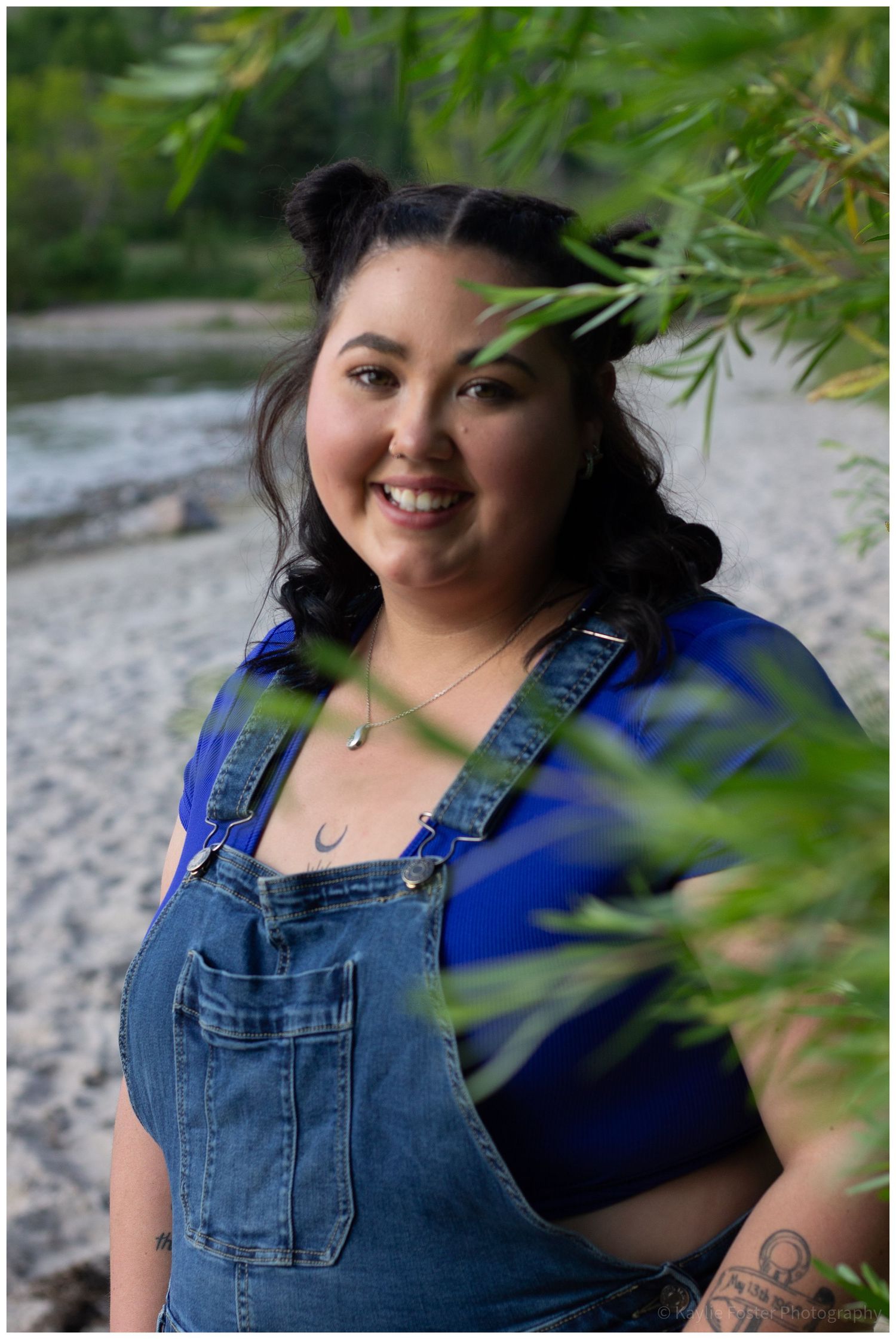 Smiling young woman in denim overalls and blue shirt poses outdoors near a river surrounded by green leaves.
