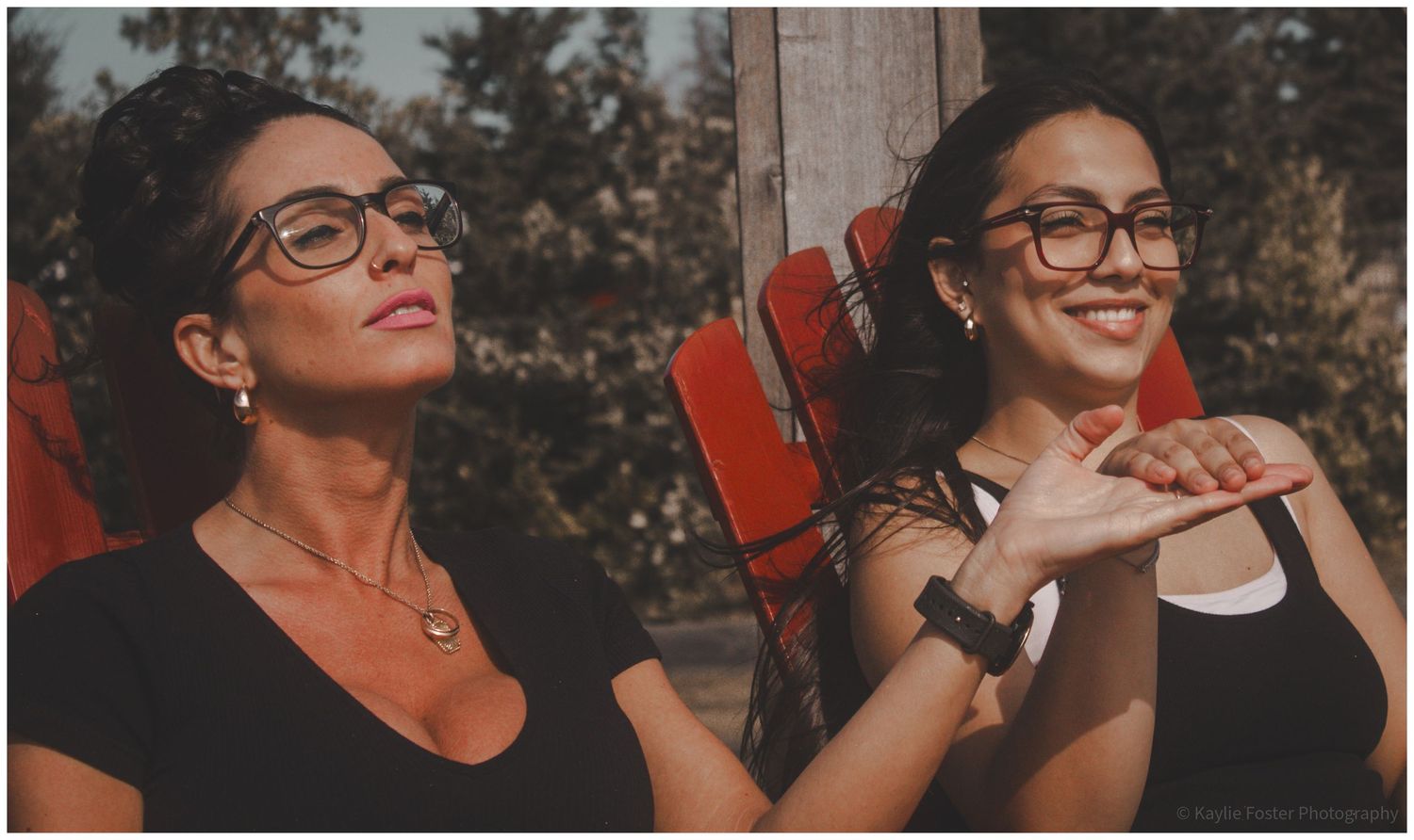 Two women wearing glasses and black tops smiling outdoors, seated in red chairs on a sunny day.