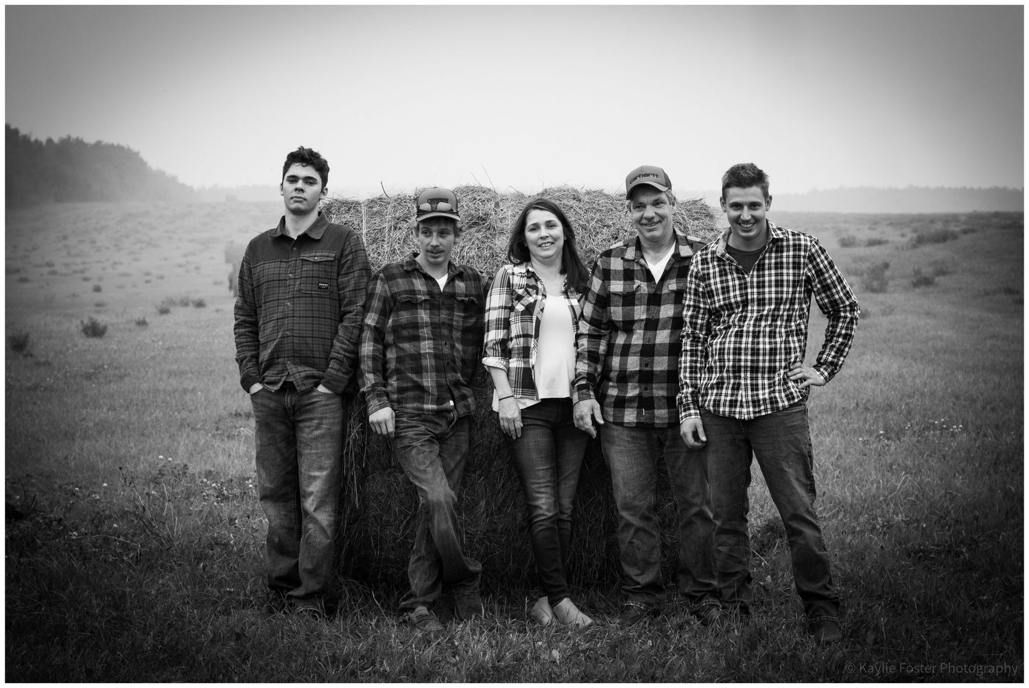 Black and white photo of four men and one woman posing by a hay bale in an open field.
