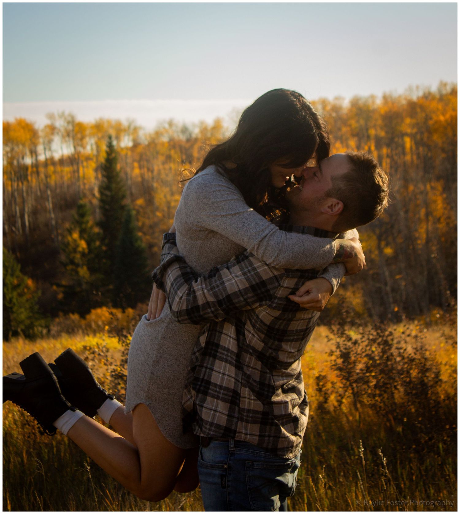 Couple sharing a romantic kiss in a golden autumn field surrounded by colorful fall foliage and evergreen trees.