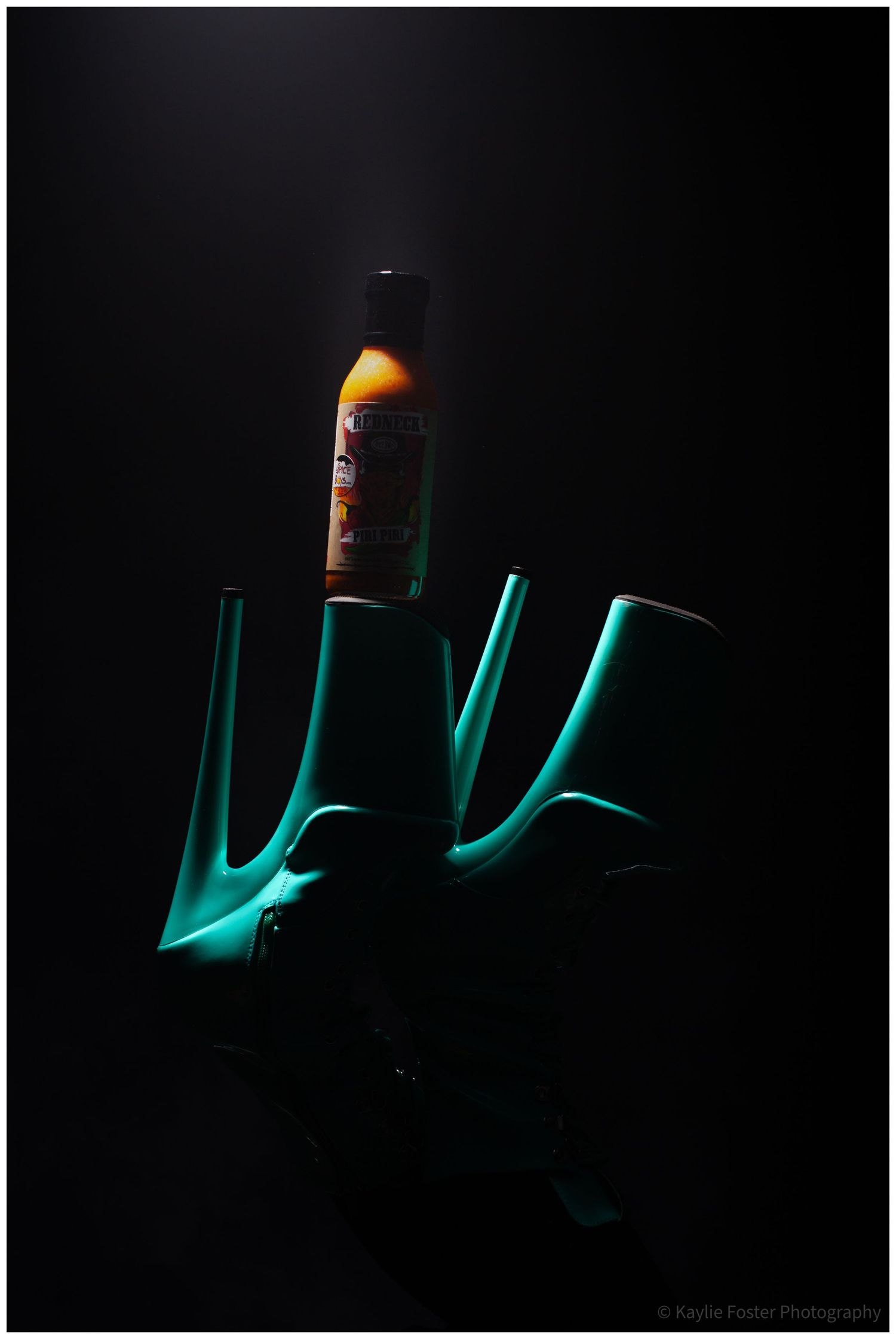 Teal platform boots holding a hot sauce bottle dramatically lit against a dark black background.