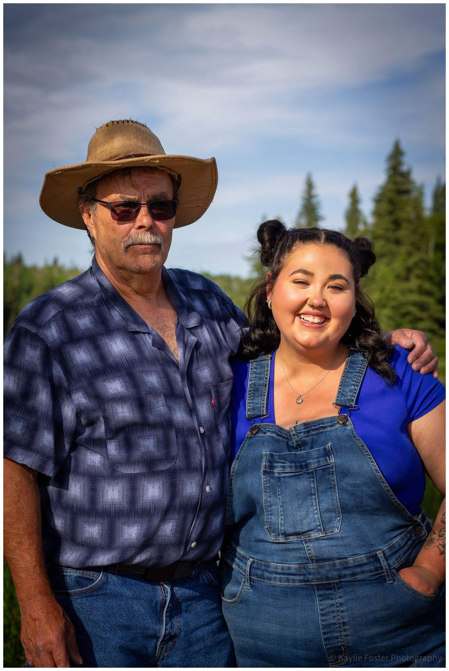Older man in cowboy hat and younger woman in denim overalls smile together outdoors.
