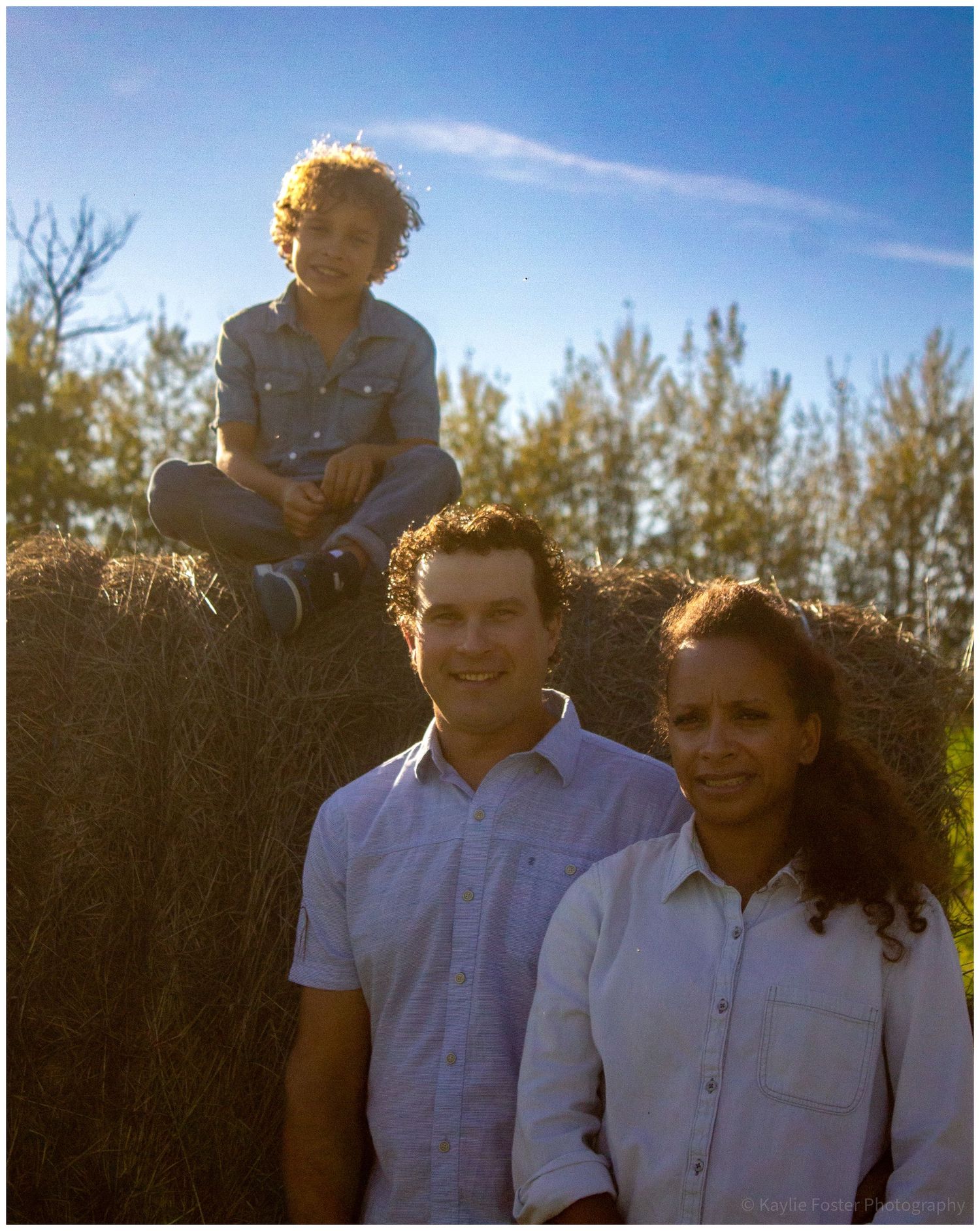 Family of three posing by hay bales under blue sky on sunny day.