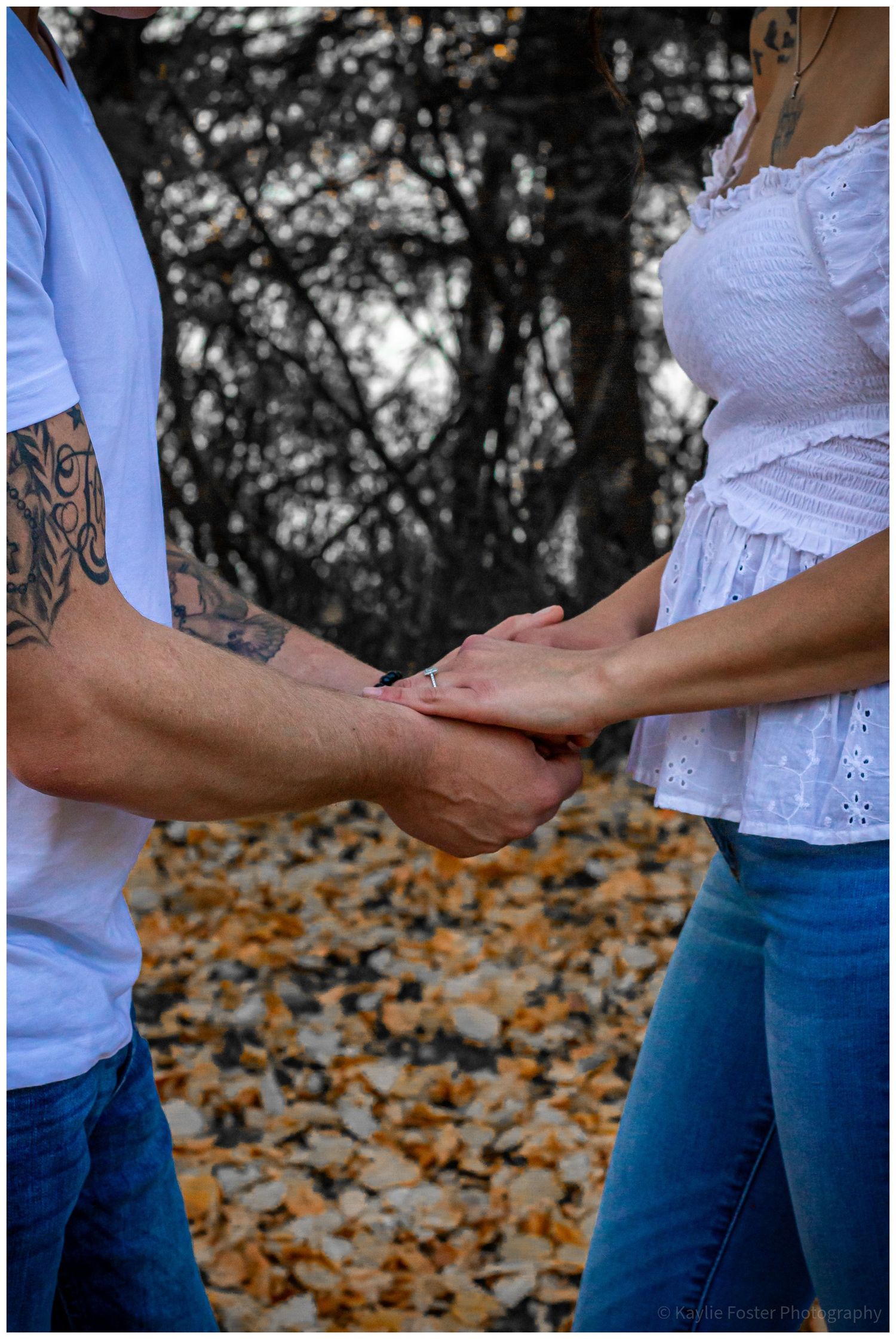 Couple holding hands outdoors in autumn, man with tattoed arm wearing white shirt, woman in white top and blue jeans.