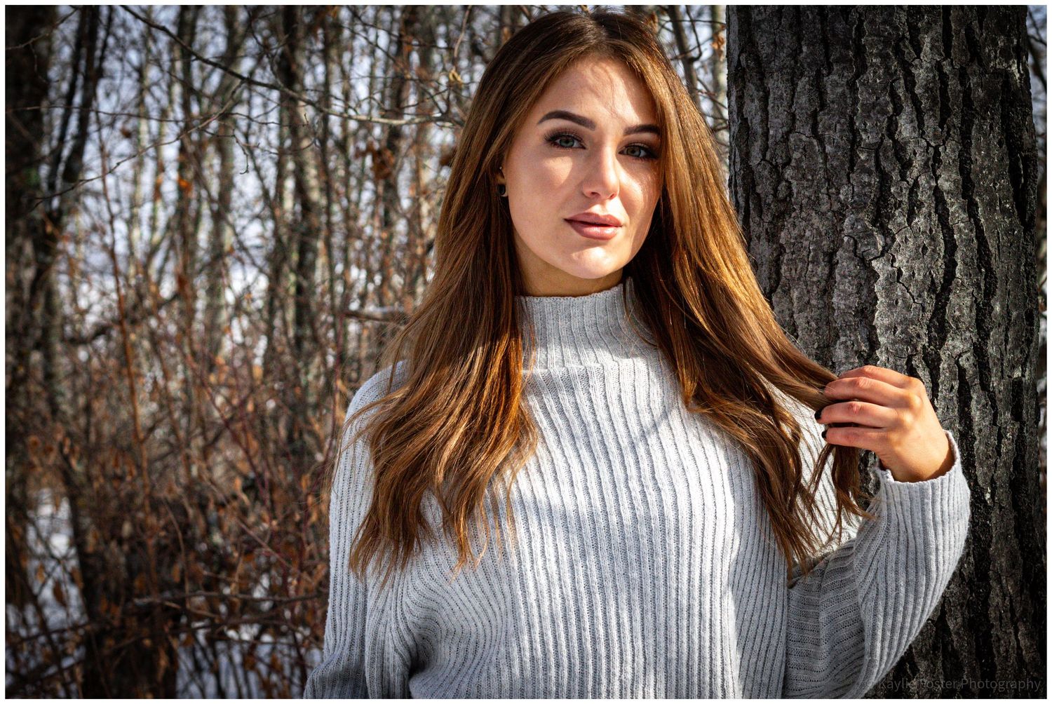 Young woman with long brown hair wearing a gray ribbed turtleneck sweater standing near a tree outdoors.