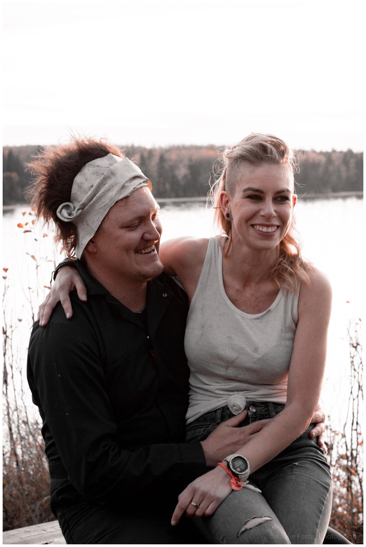 A smiling couple sitting together outdoors near a lake, the man wearing a headband and black shirt, the woman in a white tank top.