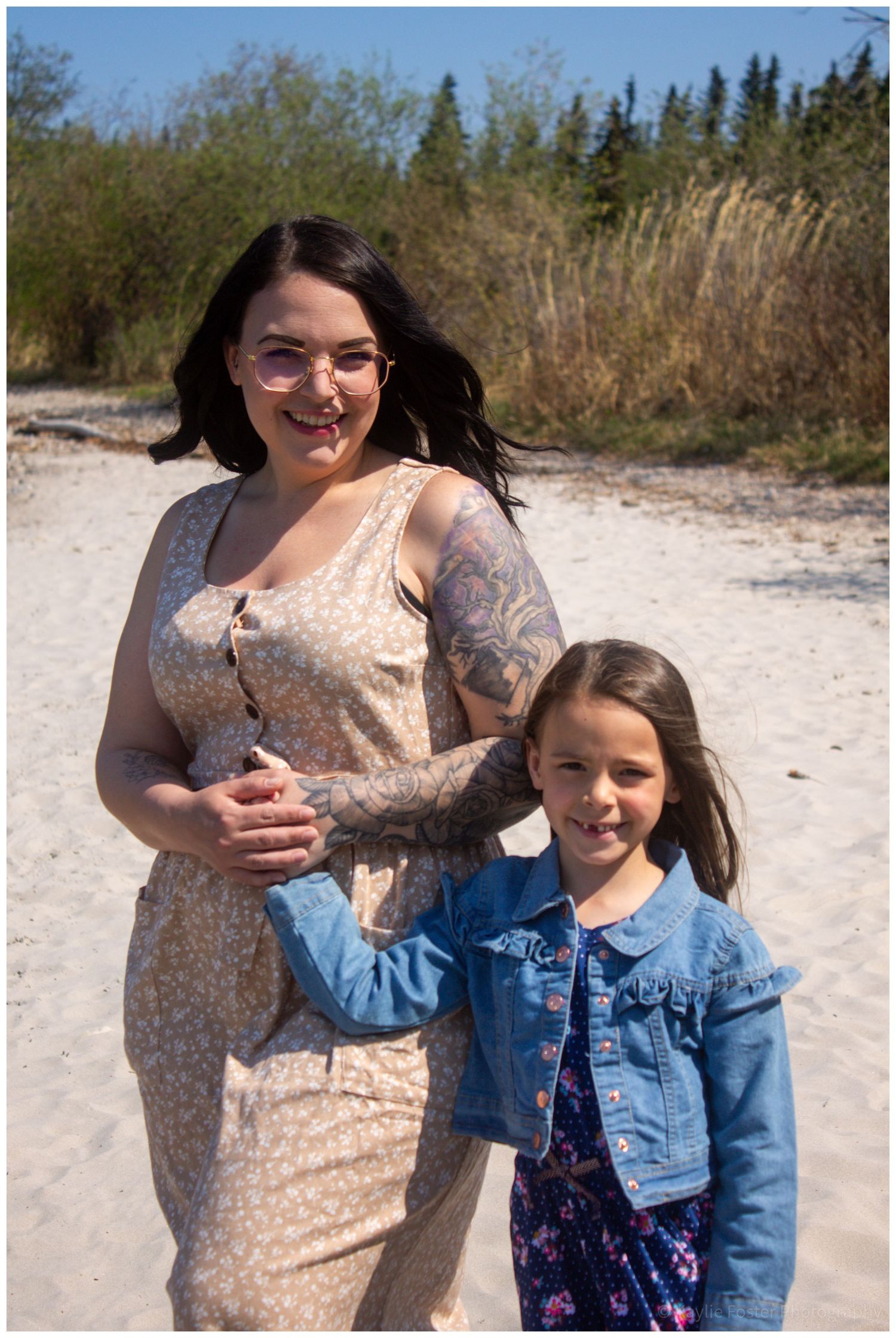 A tattooed woman in a floral dress poses with a young girl in a denim jacket on a sandy beach.