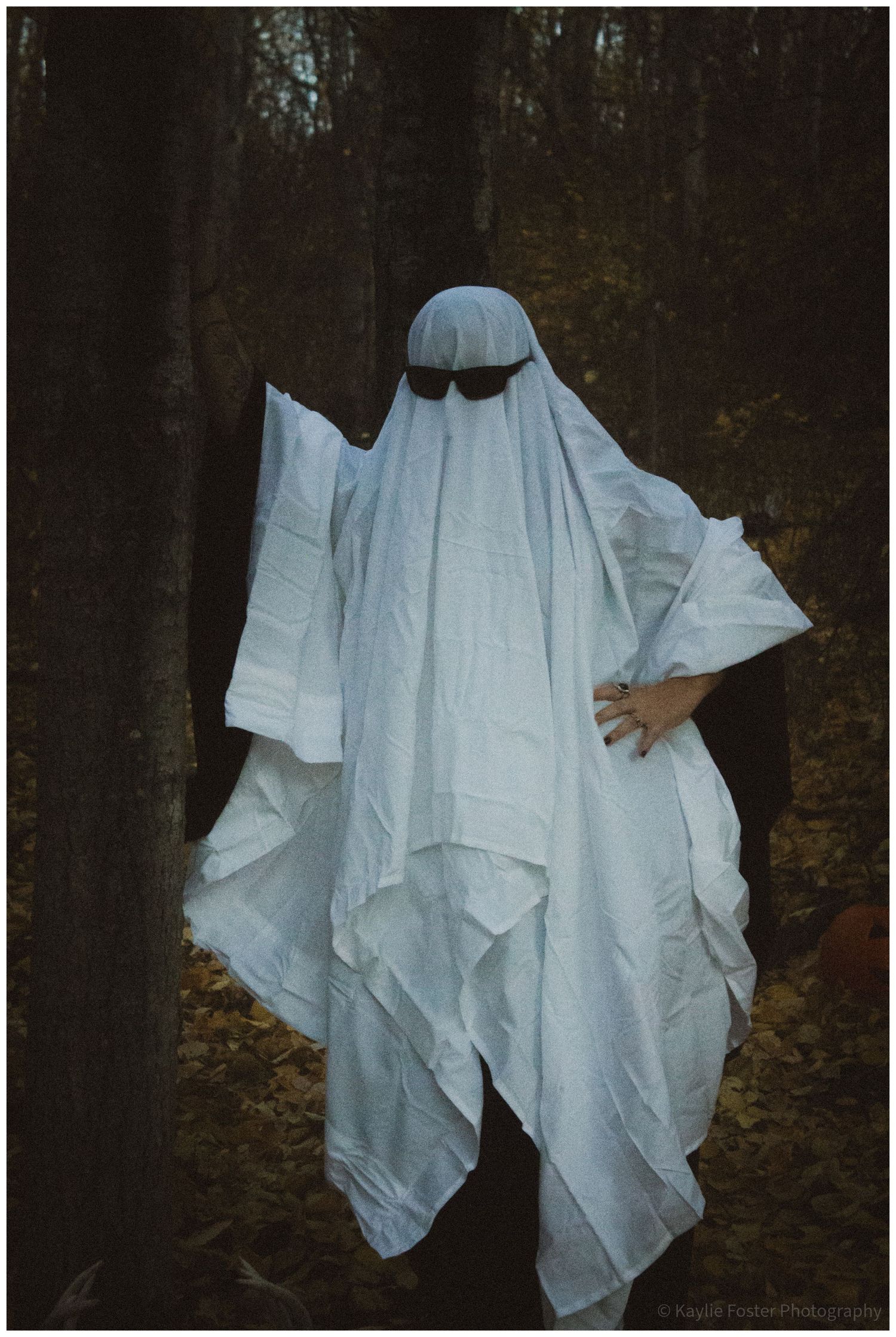 Person dressed as a ghost in white sheet with sunglasses standing in autumn forest for Halloween.