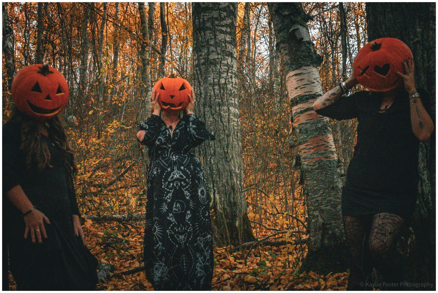 Three people holding carved pumpkins over their heads in a colorful autumn forest setting.