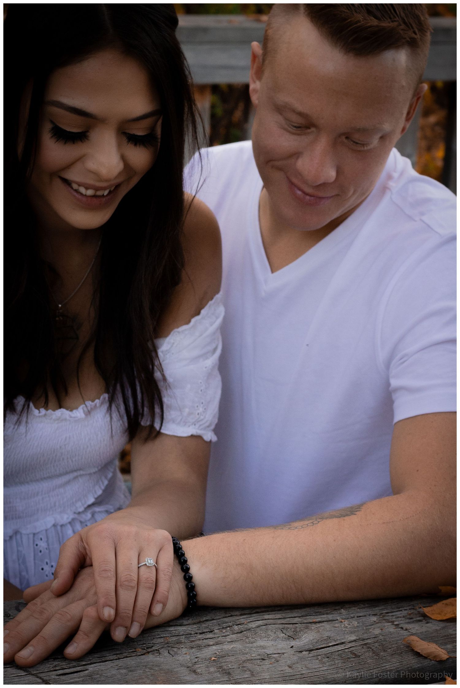 Couple smiling and admiring an engagement ring on the woman's hand while sitting at a wooden table outdoors.