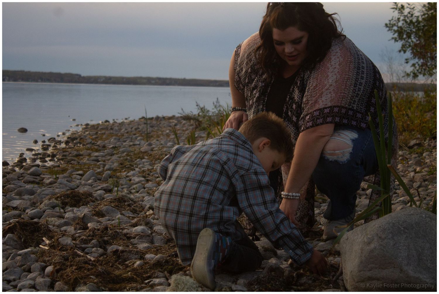Woman and child exploring rocky shoreline near calm water at dusk.
