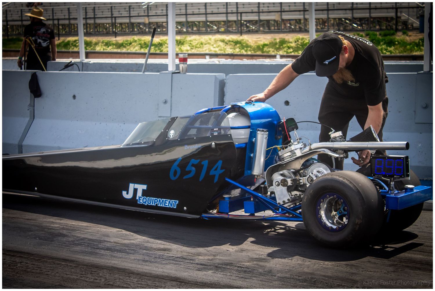Mechanic preparing blue JT dragster number 6514 on sunny drag racing track.