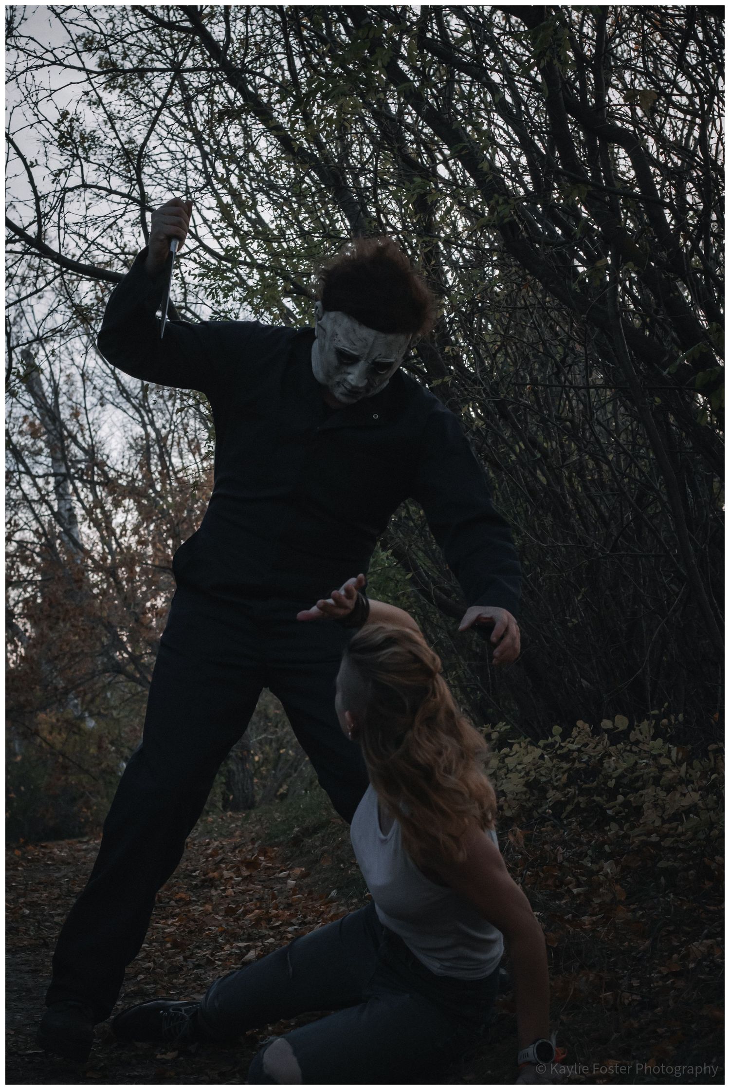 Person in Michael Myers Halloween costume mask attacking a woman in a dark forest setting.