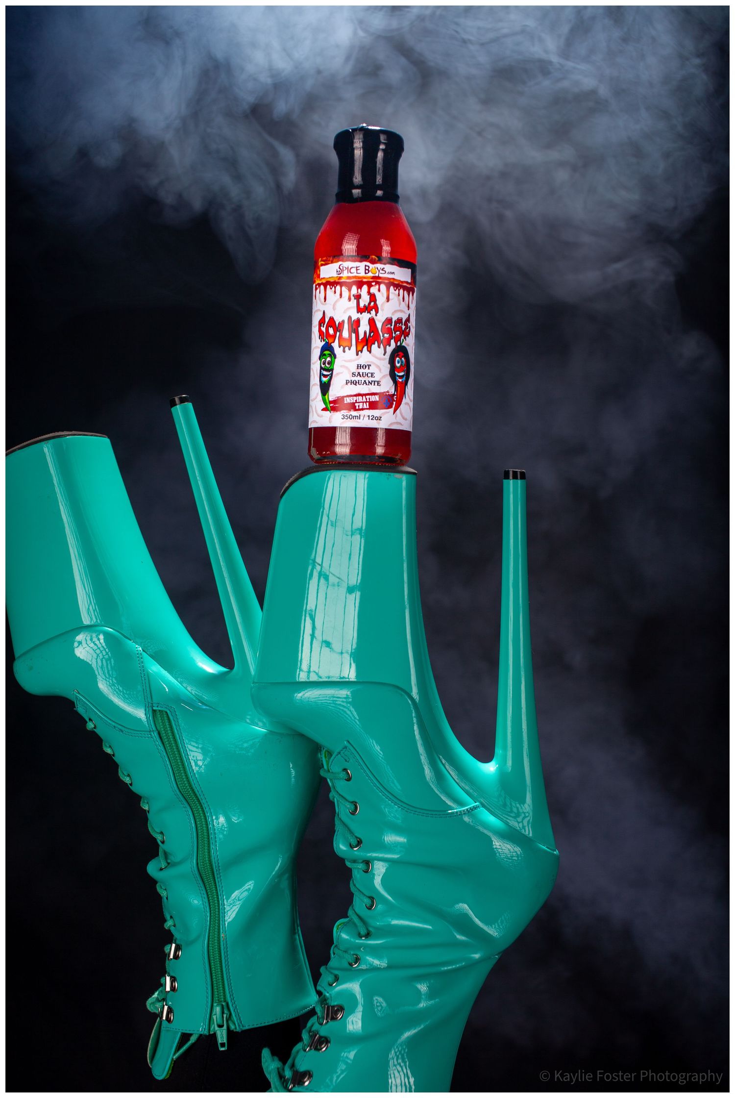 Teal platform boots balancing a red hot sauce bottle against a dramatic smoky dark background.