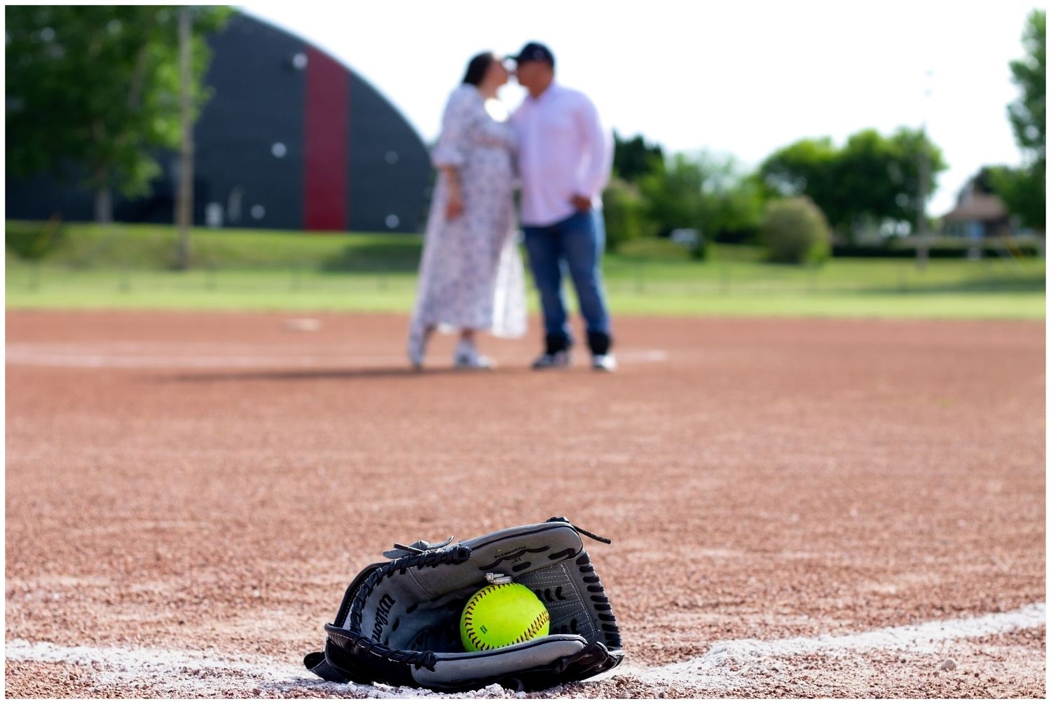 Couple sharing a kiss on a softball field with a glove and yellow softball in the foreground.