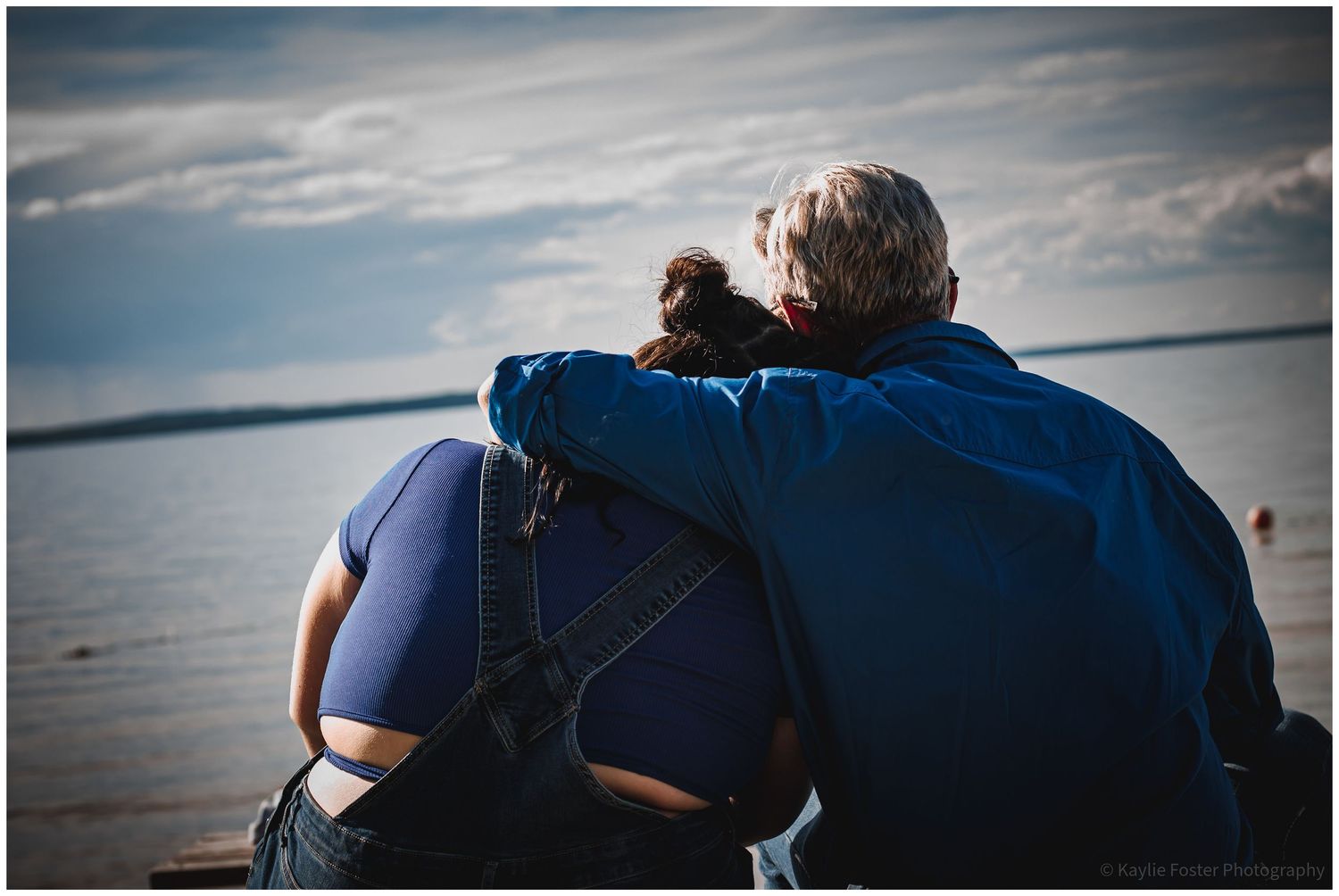 Elderly couple sitting together by the water, man with arm around woman, sharing a tender moment overlooking a calm sea.