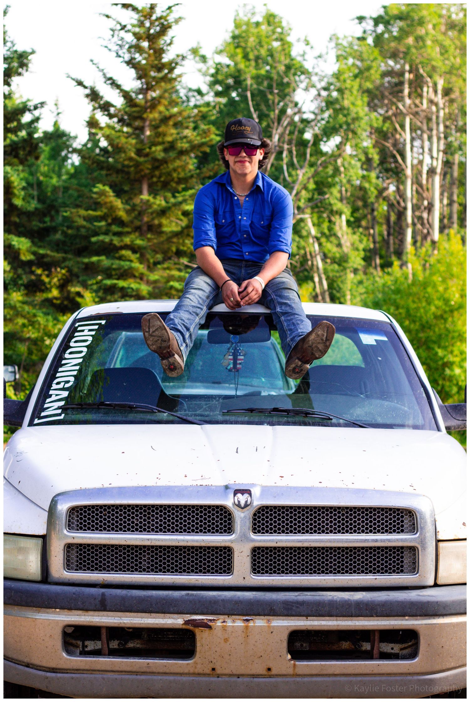 Teen boy in blue shirt and bucket hat sits on hood of muddy Dodge Ram truck outdoors.