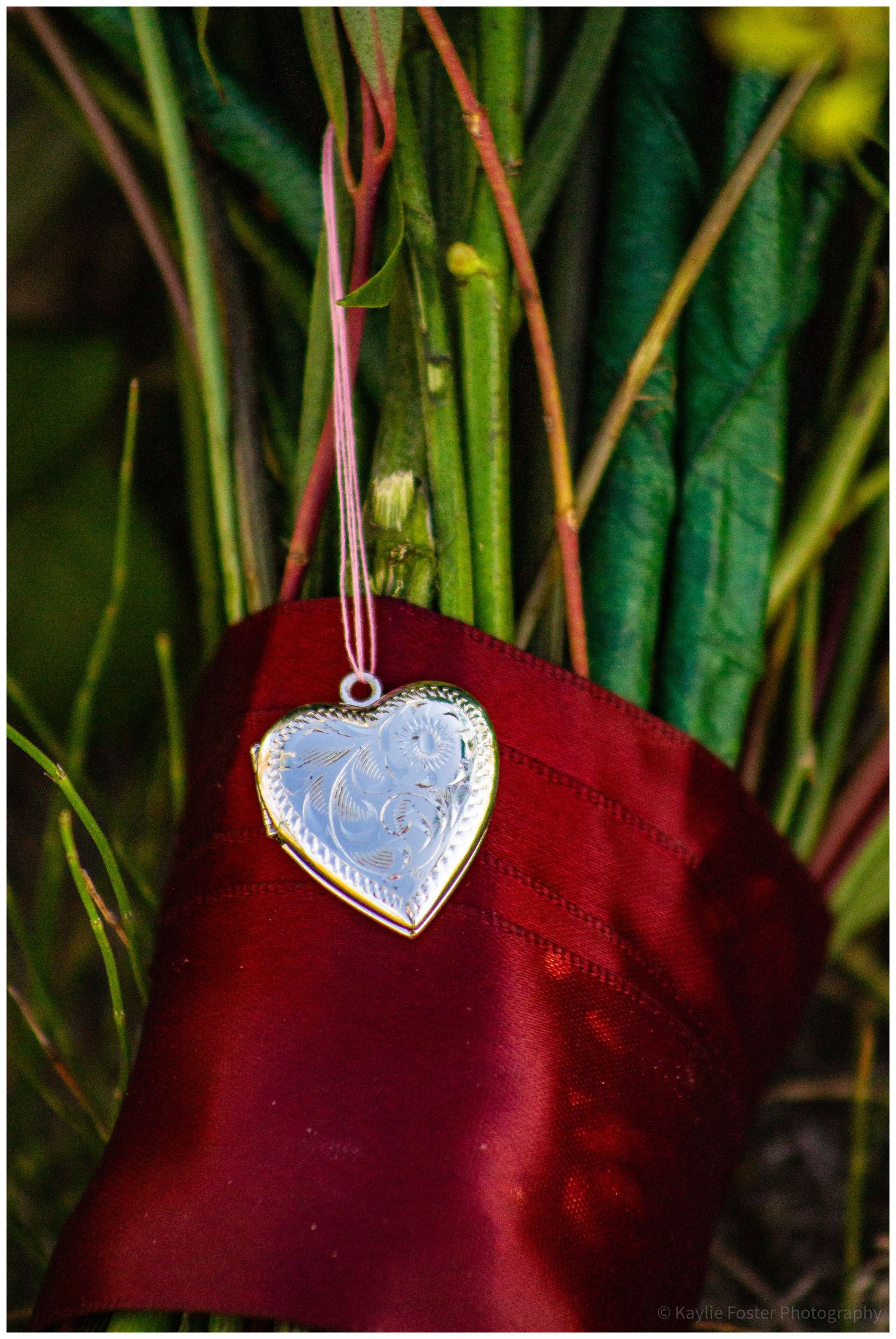 Silver heart-shaped locket pendant hanging on pink ribbon against red satin fabric and green plants backdrop.