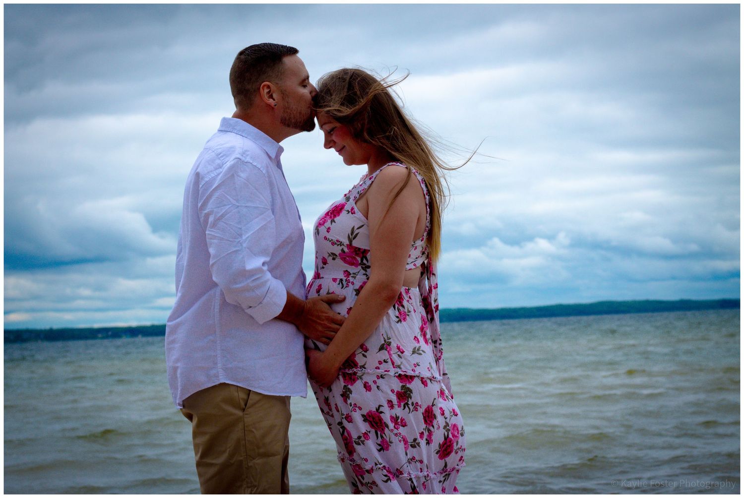Couple sharing a tender moment on a beach, woman in floral dress with baby bump, man in white shirt, cloudy sky.