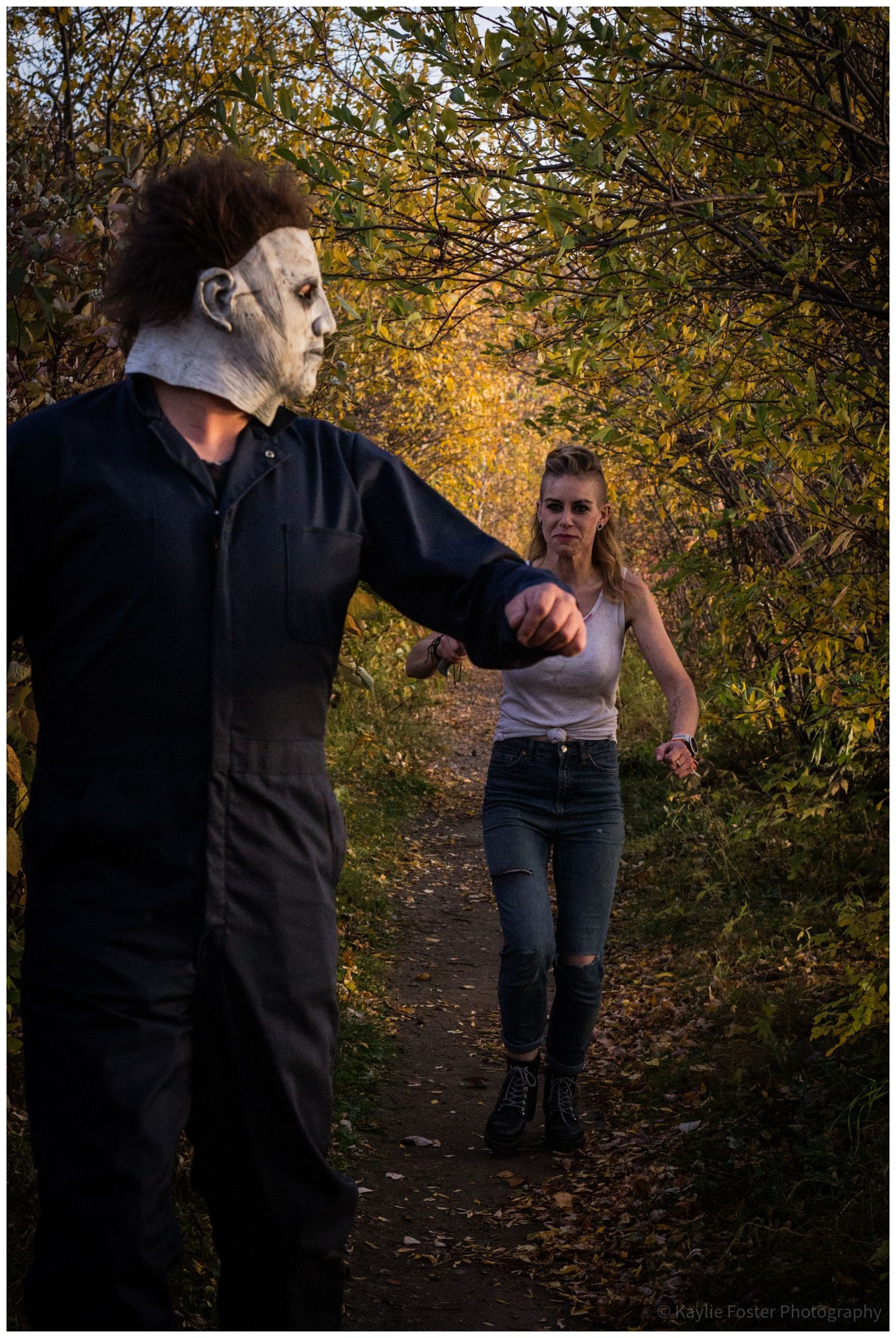 Michael Myers Halloween costume chasing woman on forest path in fall foliage.