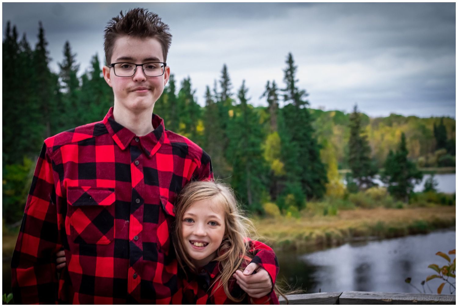 Two siblings in matching red plaid shirts pose outdoors near a forest lake on a cloudy day.