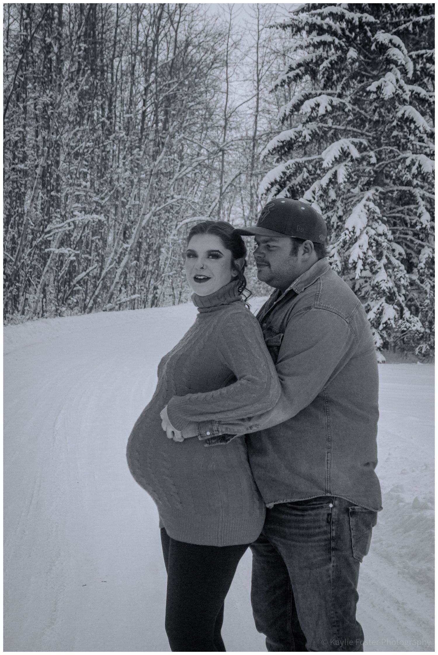 Pregnant couple embracing on snowy winter road surrounded by snow-covered trees in black and white maternity photo.