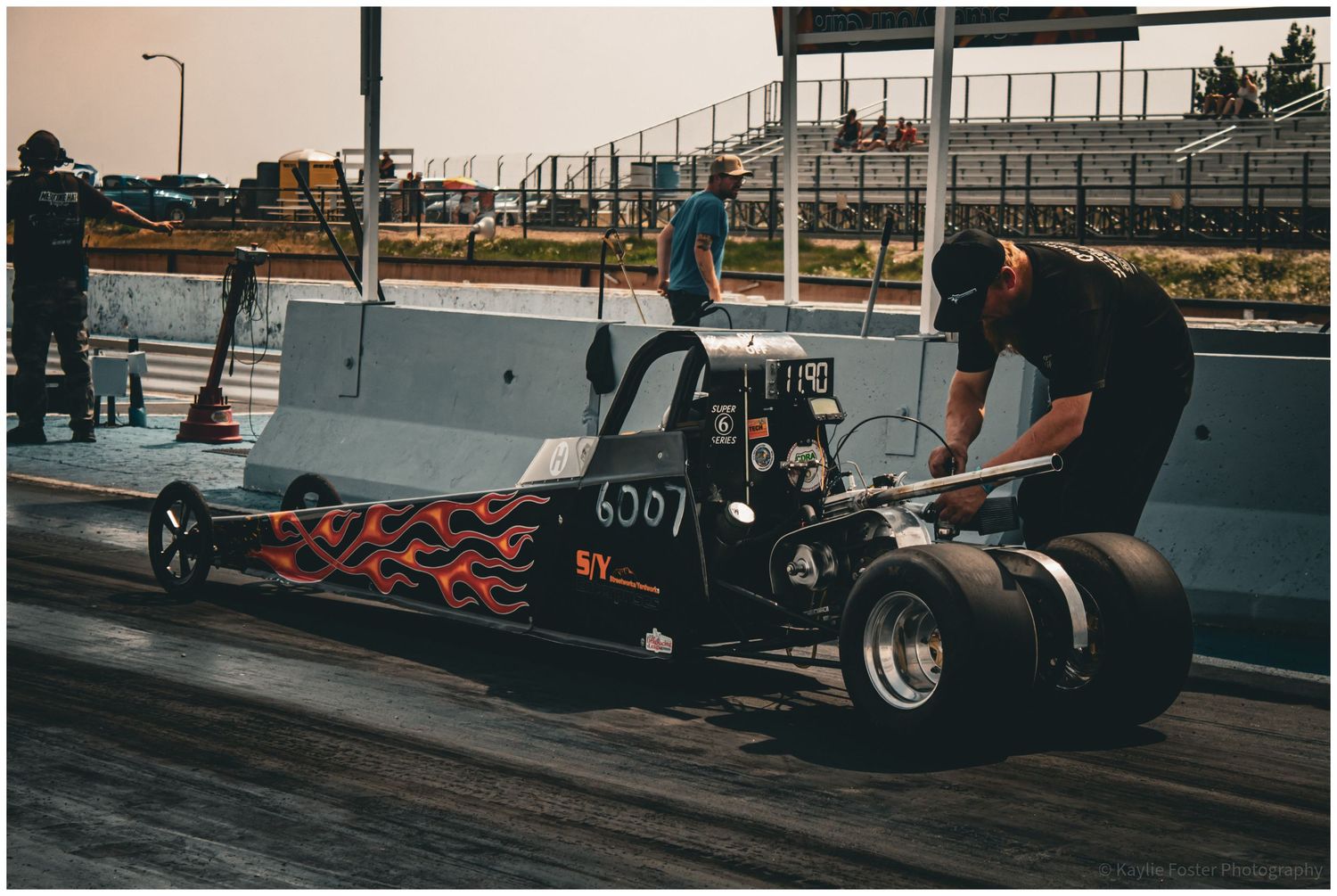 Black flame dragster number 6007 on drag strip being inspected by crew member at sunset.