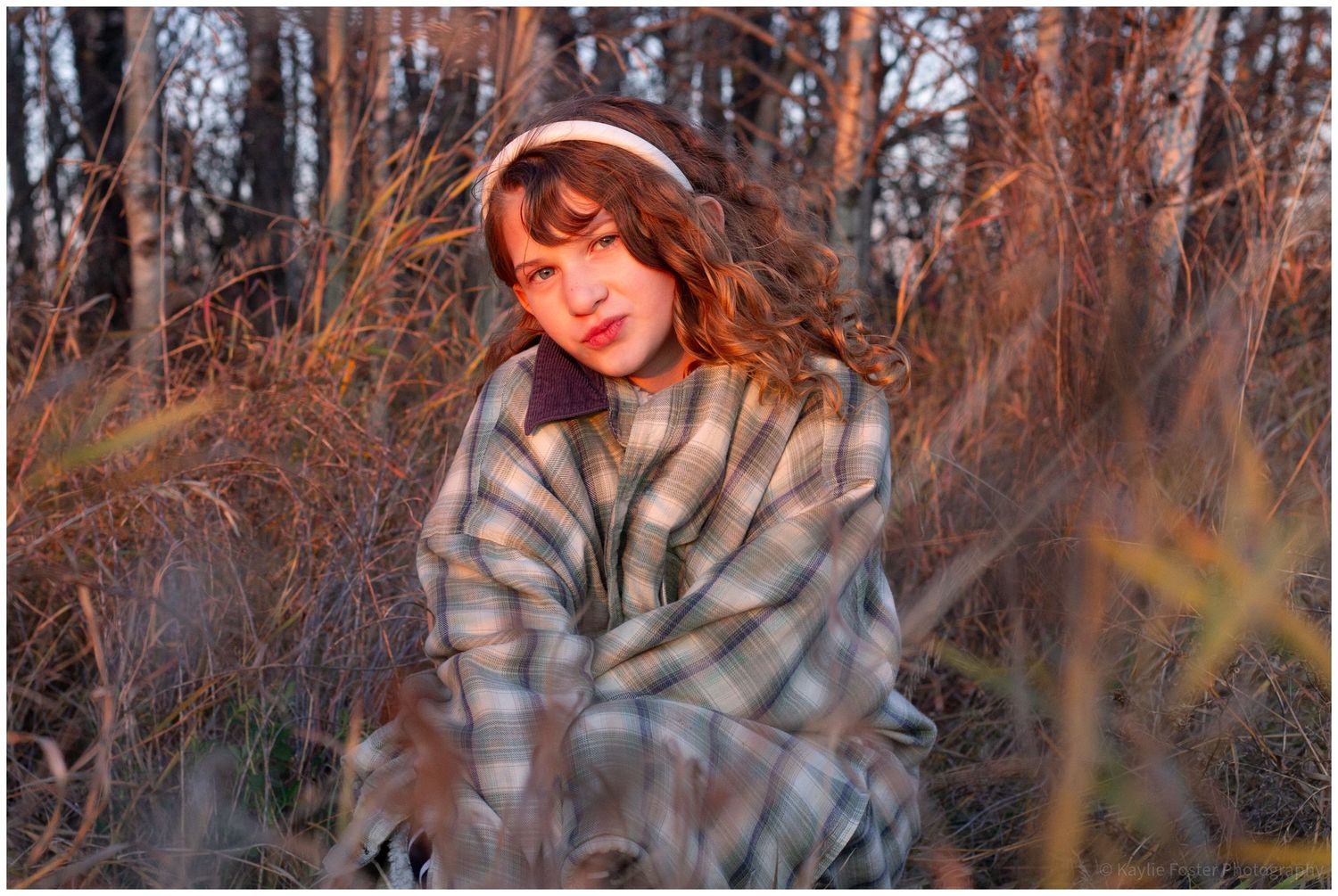 Young woman in plaid coat and white earmuffs sitting among tall dry grasses in soft golden hour light.
