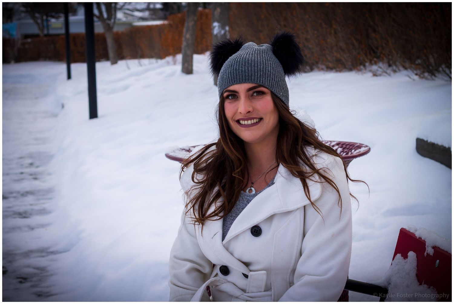 Young woman in white coat and gray pom-pom beanie smiling outdoors in snowy winter setting.