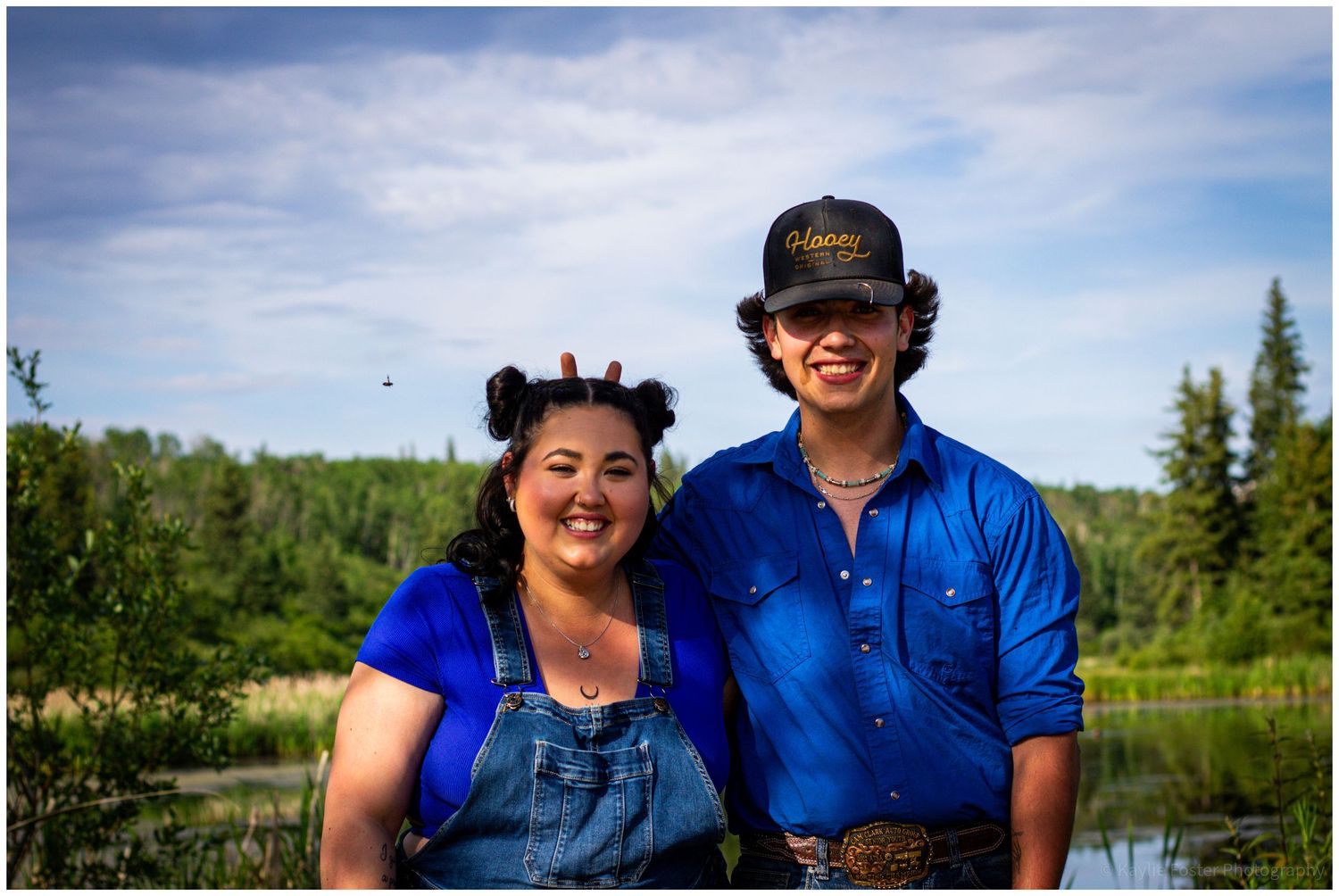 A smiling couple poses outdoors near a lake surrounded by lush green trees under a partly cloudy sky.