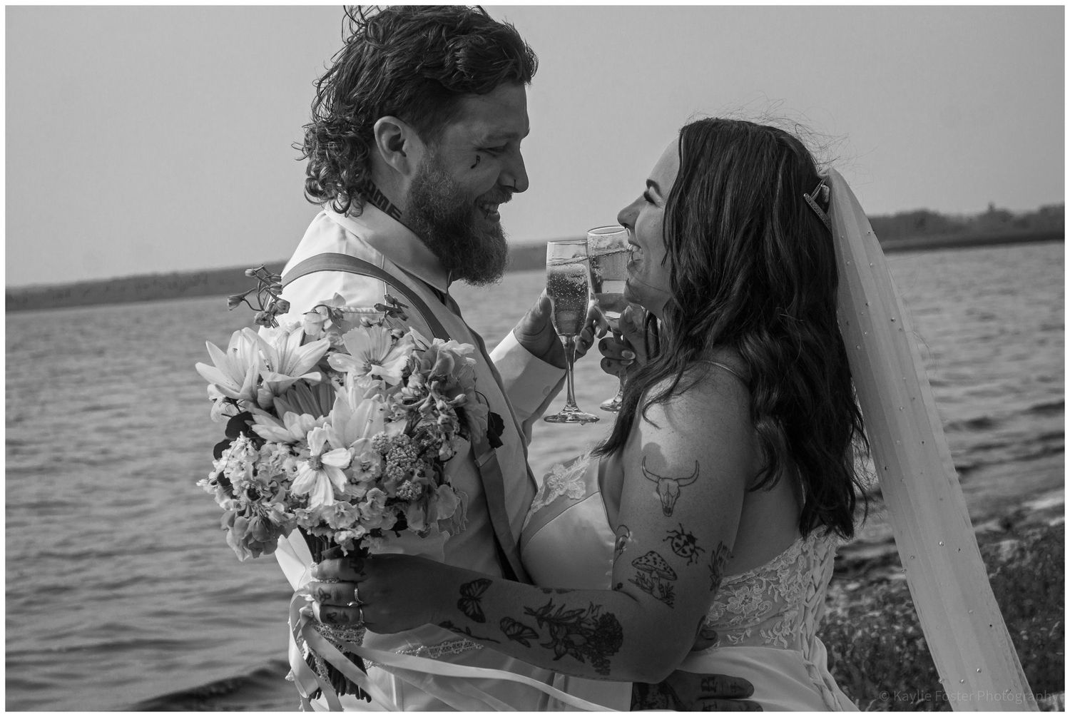 Black and white photo of a bearded groom and veiled bride toasting with champagne near a waterfront.